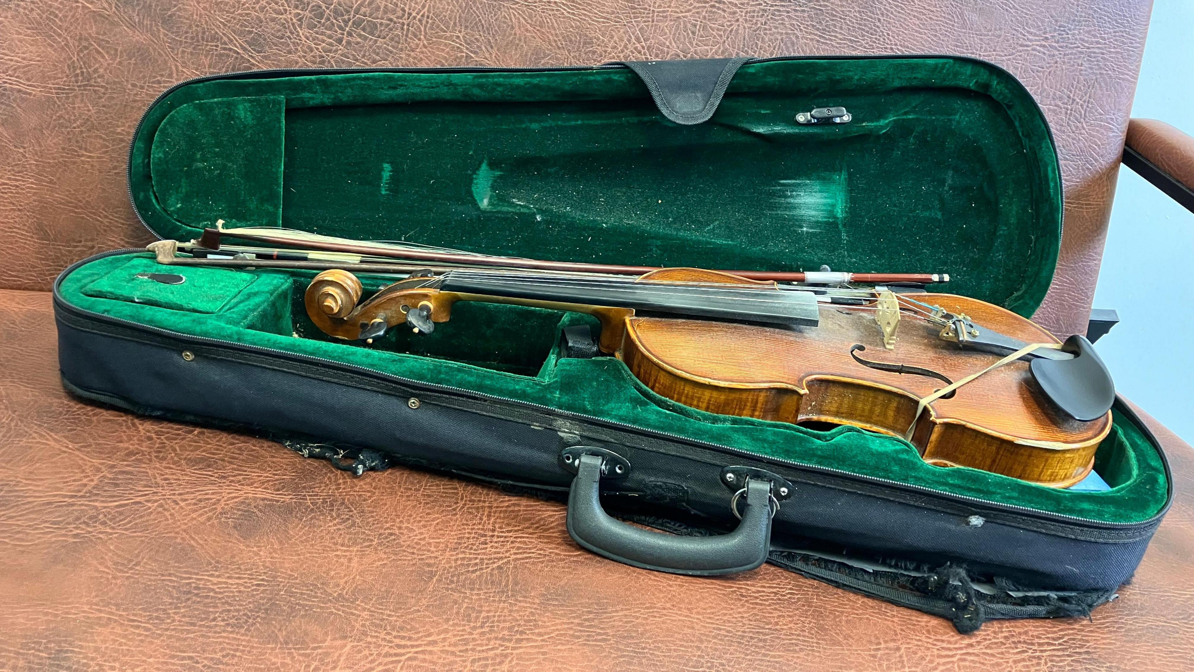 A black violin case with a green velvet inside holding a violin and two broken bows. The case is very frayed, ripped and the handle has broken off. 