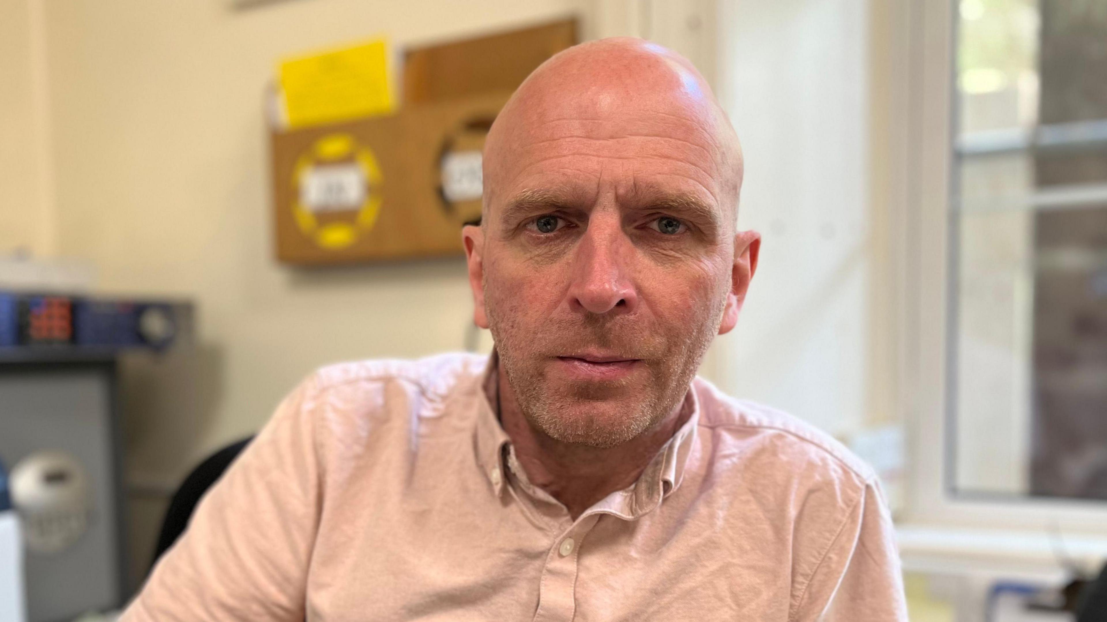 Scott Jeffery in a long sleeved pale pink shirt open and the collar sitting on an office chair looking directly at the camera with a neutral expression