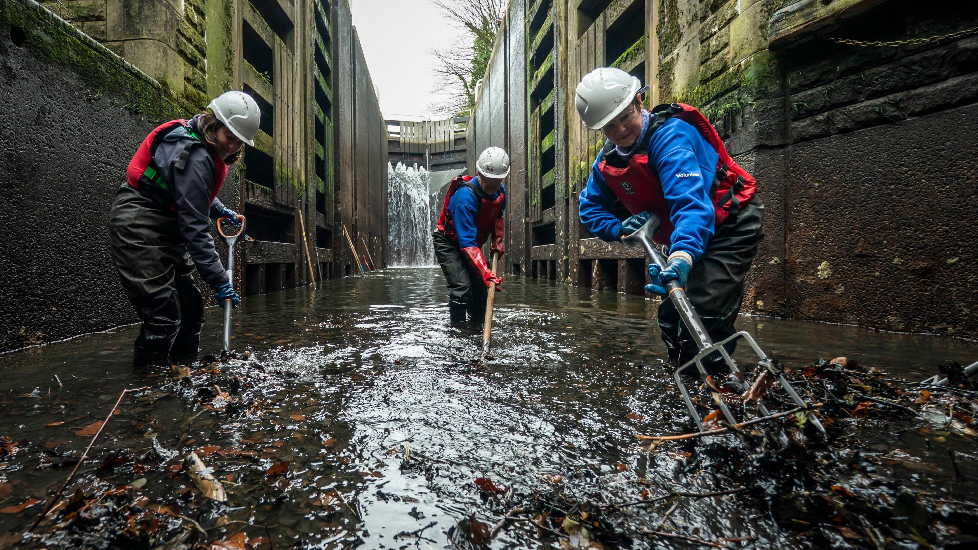 Three people wearing waterproofs rake leaves out of the canal.