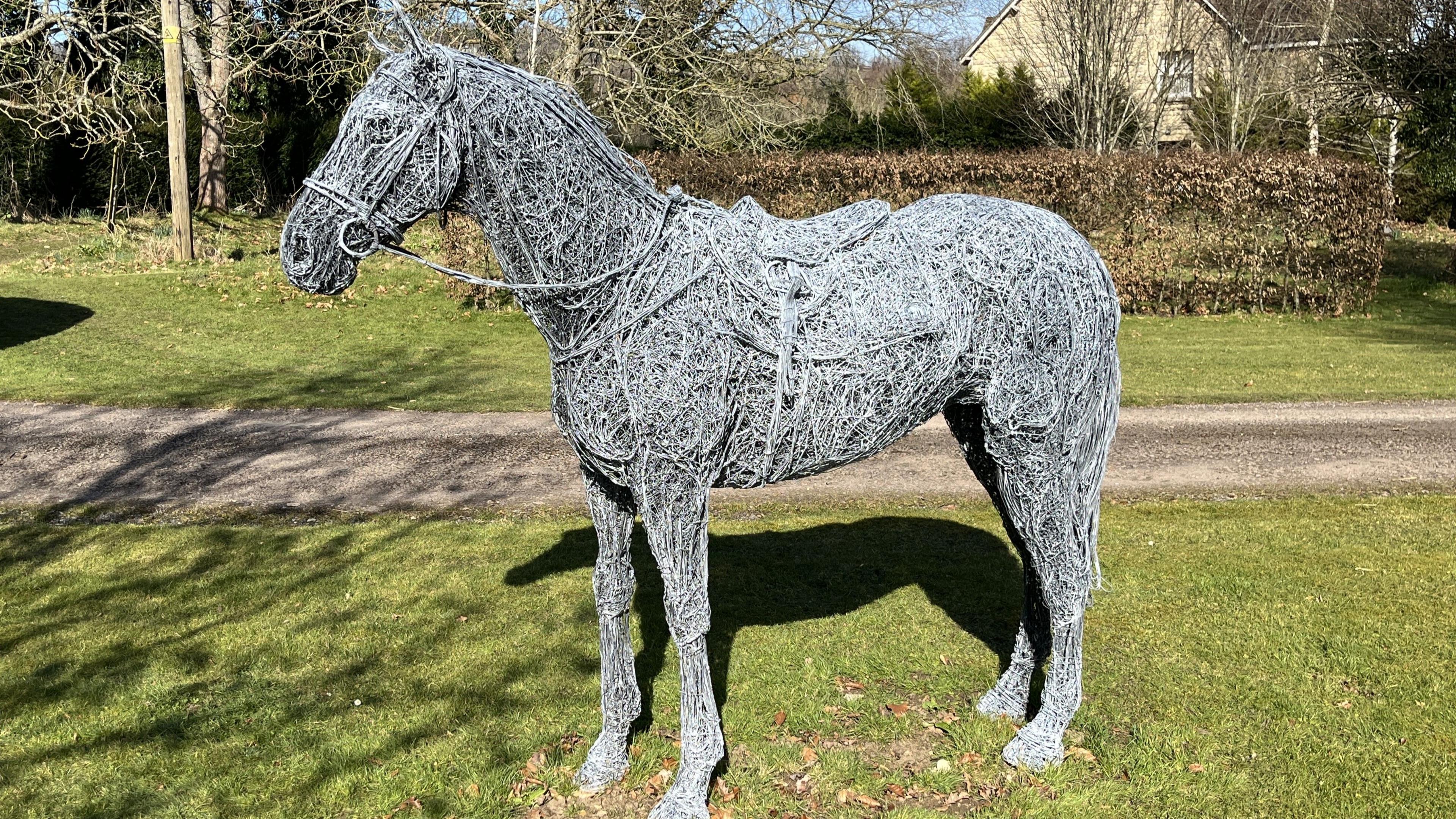 A sculpture of a horse made from galvanised grey steel. It is positioned on a patch of grass beside a gravel path, with bare trees in the background. The intricate horse sculpture is wearing a bridle, reins and a saddle.