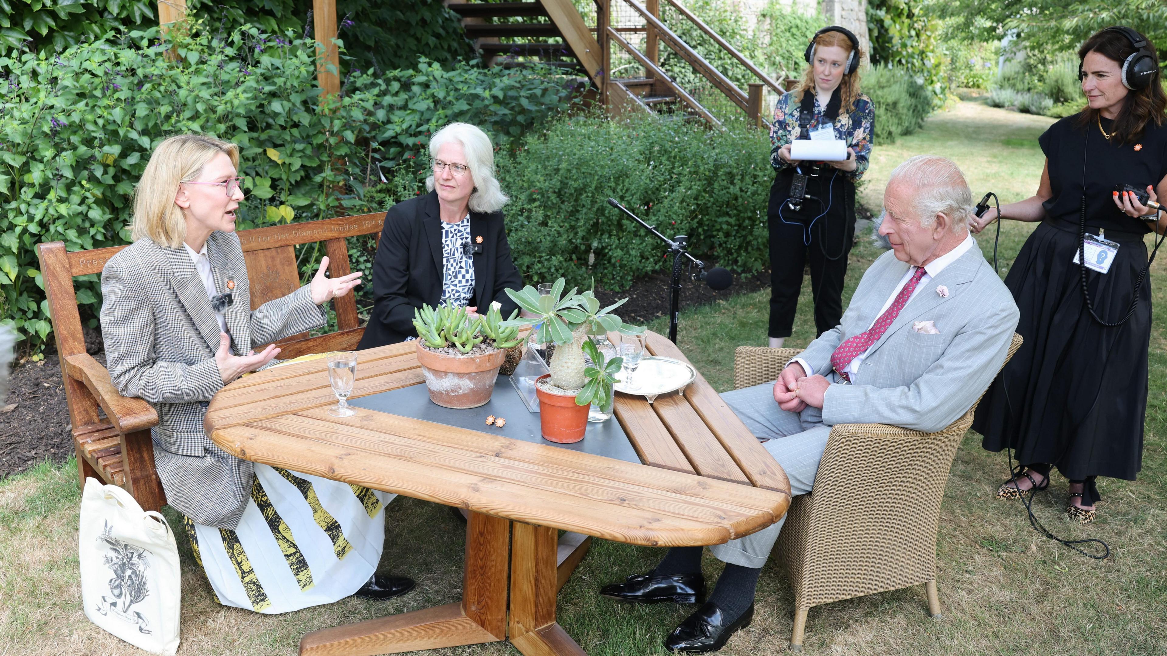 On the right of the picture is the King wearing a light grey suit and a red patterned tie sitting in a taupe garden chair with a wooden triangular table in front of him. Across the other side of the table, sitting on a wooden bench on the left side of the screen is Cate Blanchett who is gesticulating with both hands as she speaks. Next to her is Elinor Breman a scientist from Kew with shoulder length grey hair wearing a black jacket and a floral dress. There are some plants on the table and a microphone on a stand to the side. A few steps back, holing more radio equipment are the producers of the podcast.