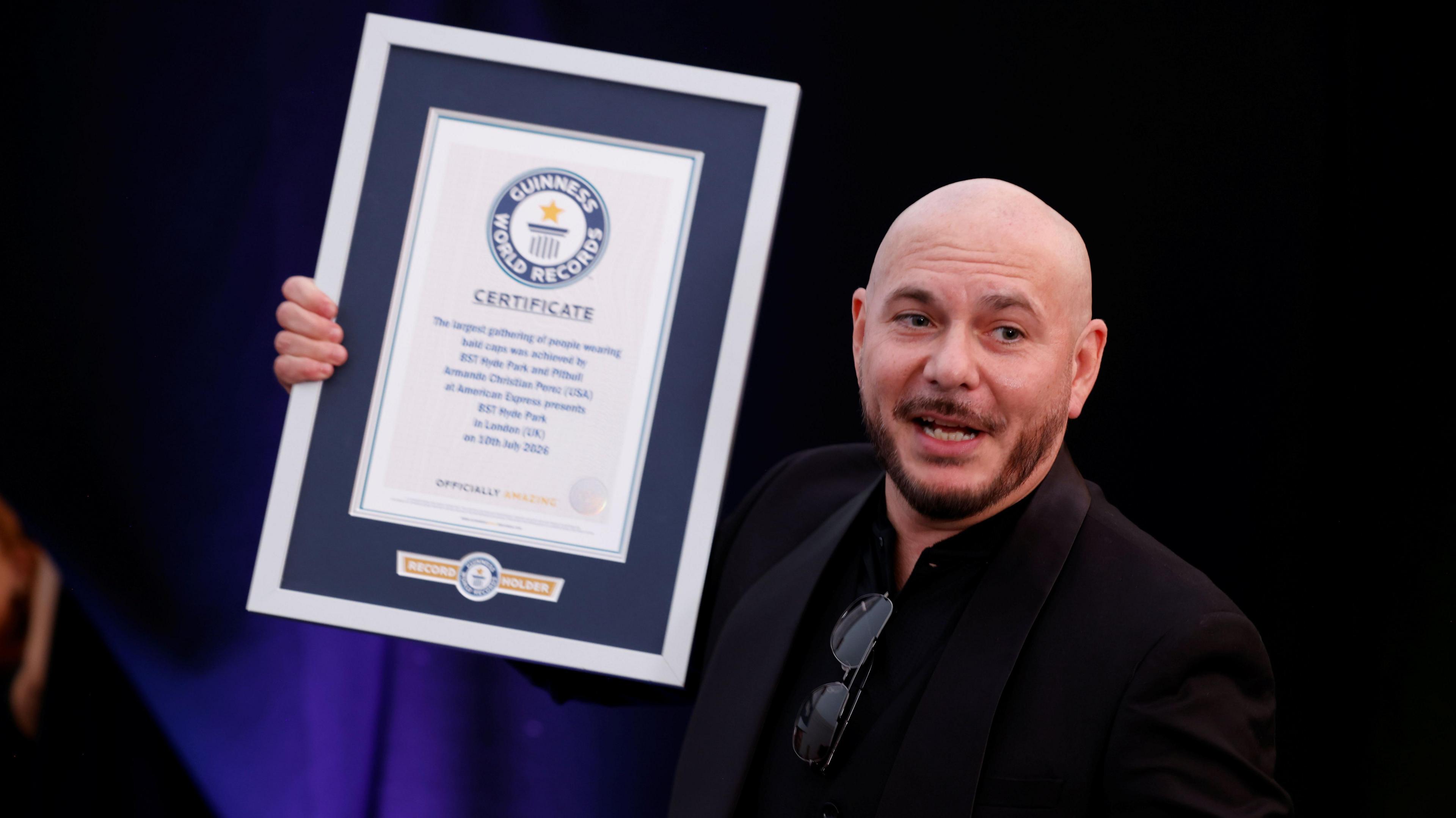 Pitbull wears a dark suit jacket over a dark shirt. He is holding a Guinness World Record award.