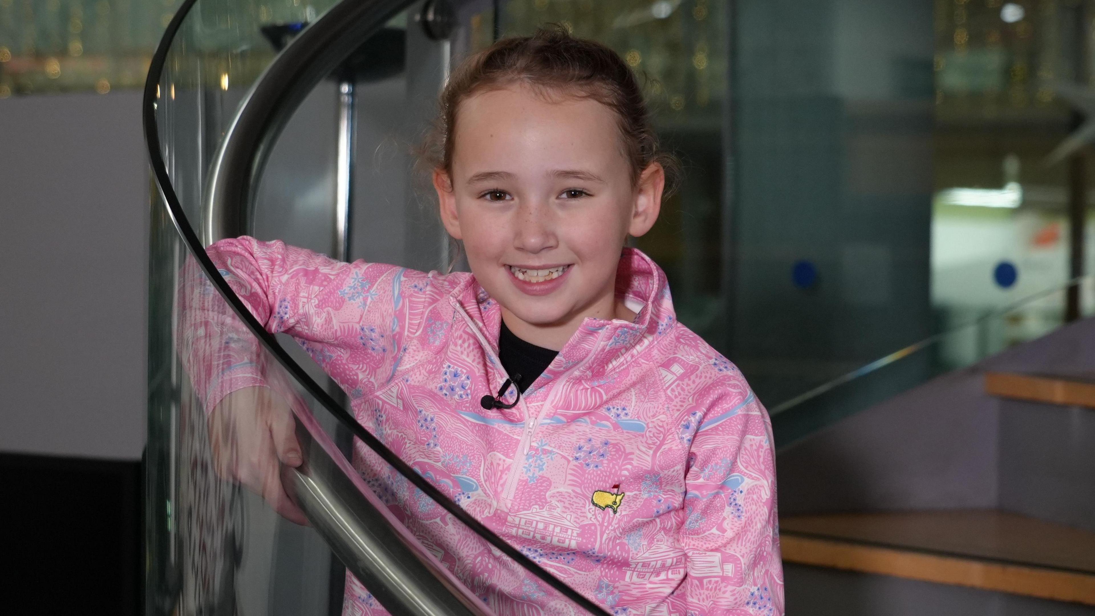 Hope is in a pink zip-up sweatshirt. She is stood on the stairs of a glass spiral staircase. She is looking at the camera and is smiling.