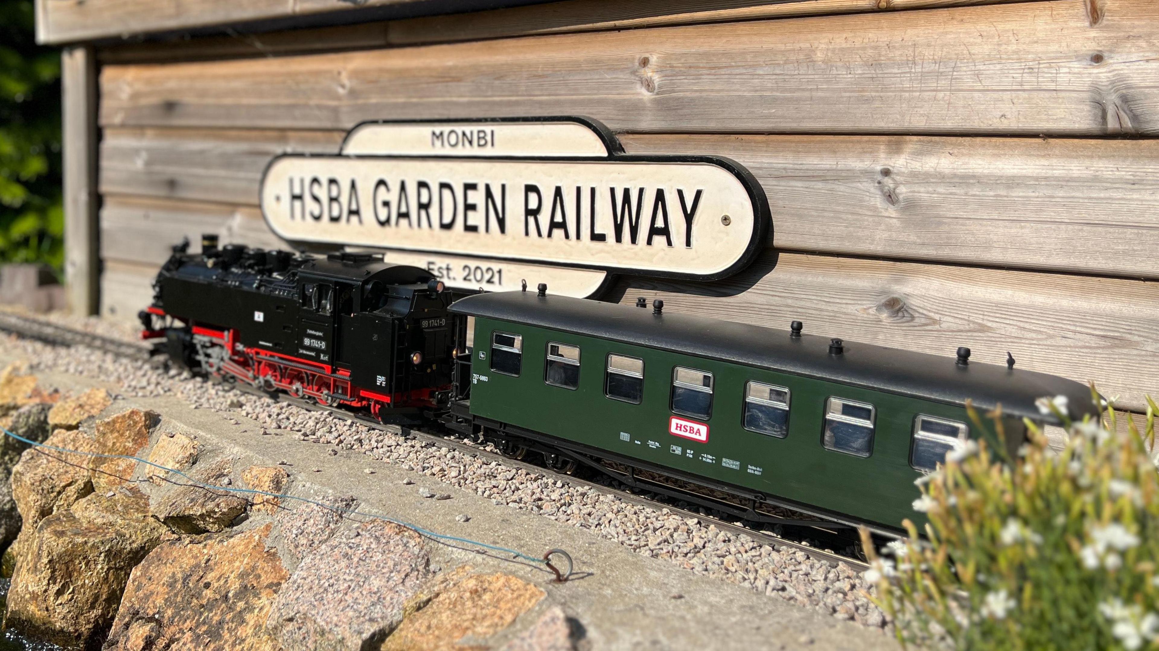 A model train is in front of a sign that says HSBA Garden Railway