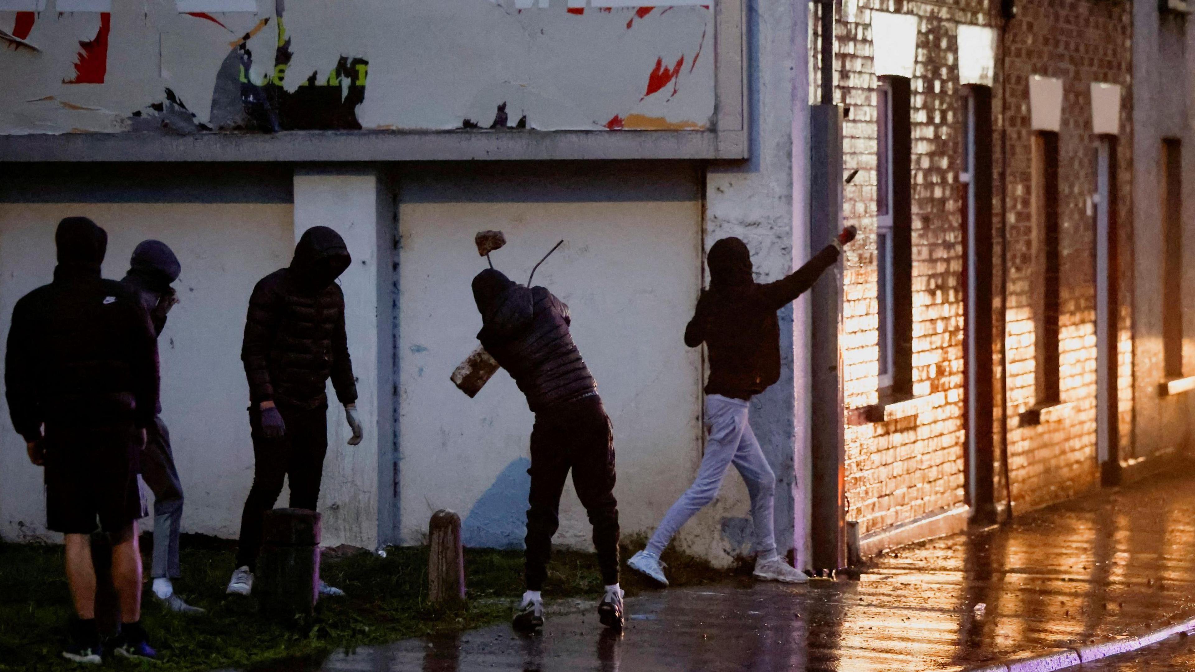 Demonstrators prepare to throw objects towards the police while they take cover behind a wall. The police is out of frame. The men are wearing black puffer coats and their faces are concealed.