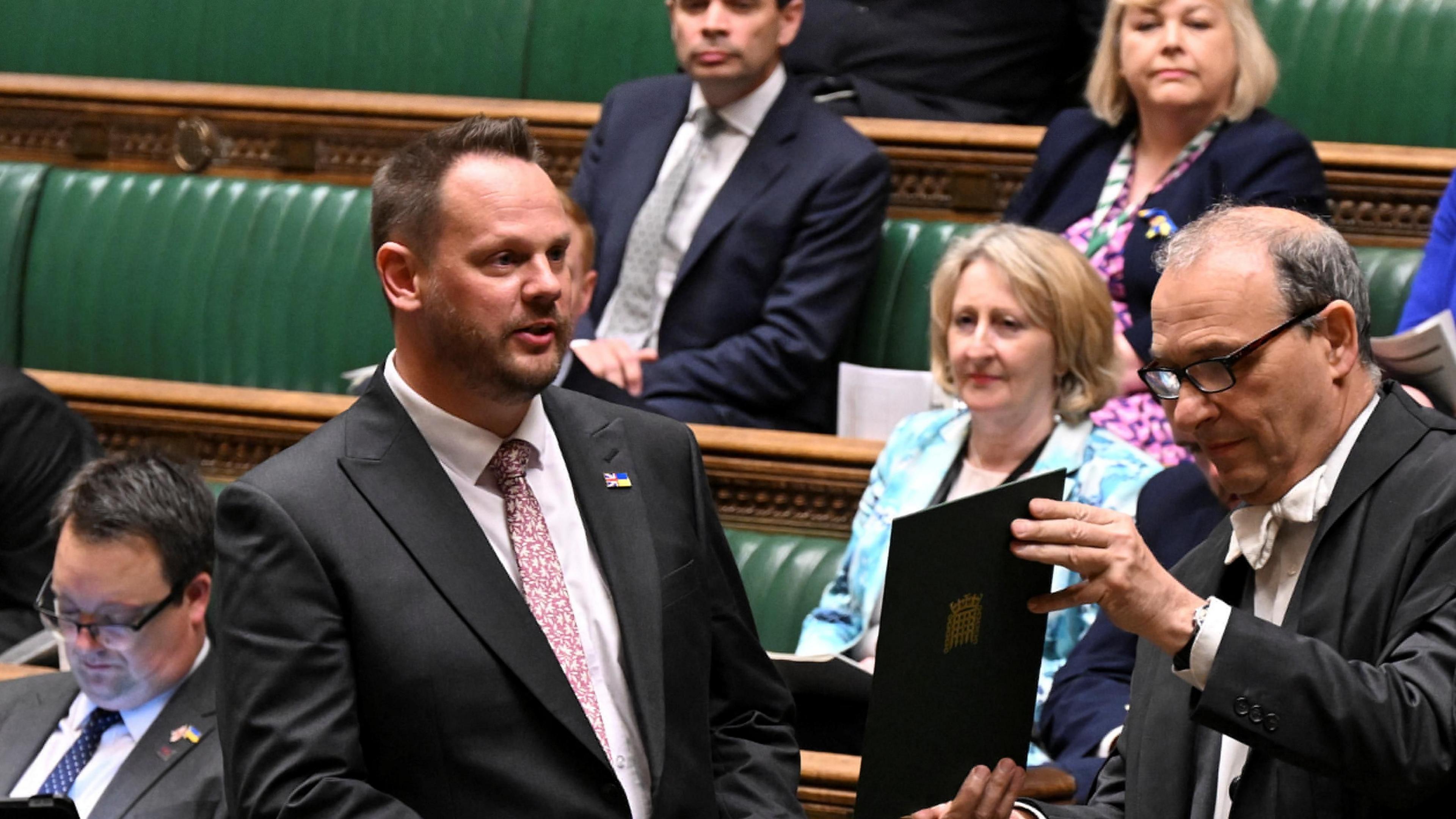 A man in a suit standing up talking in the house of commons among other MPs and the green benches