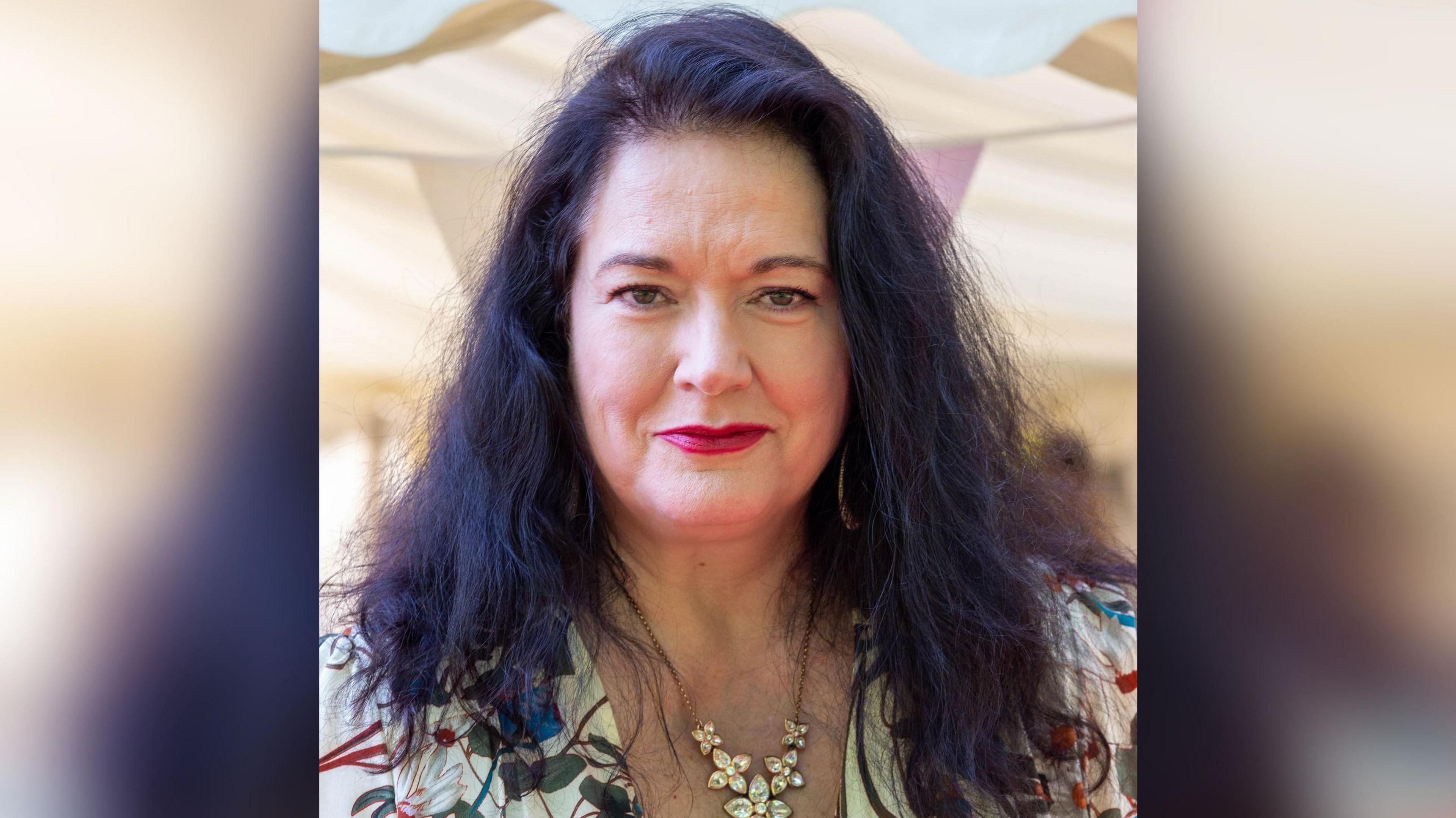 Image shows Ann Millington looking at the camera, she has long dark hair and is wearing a floral top