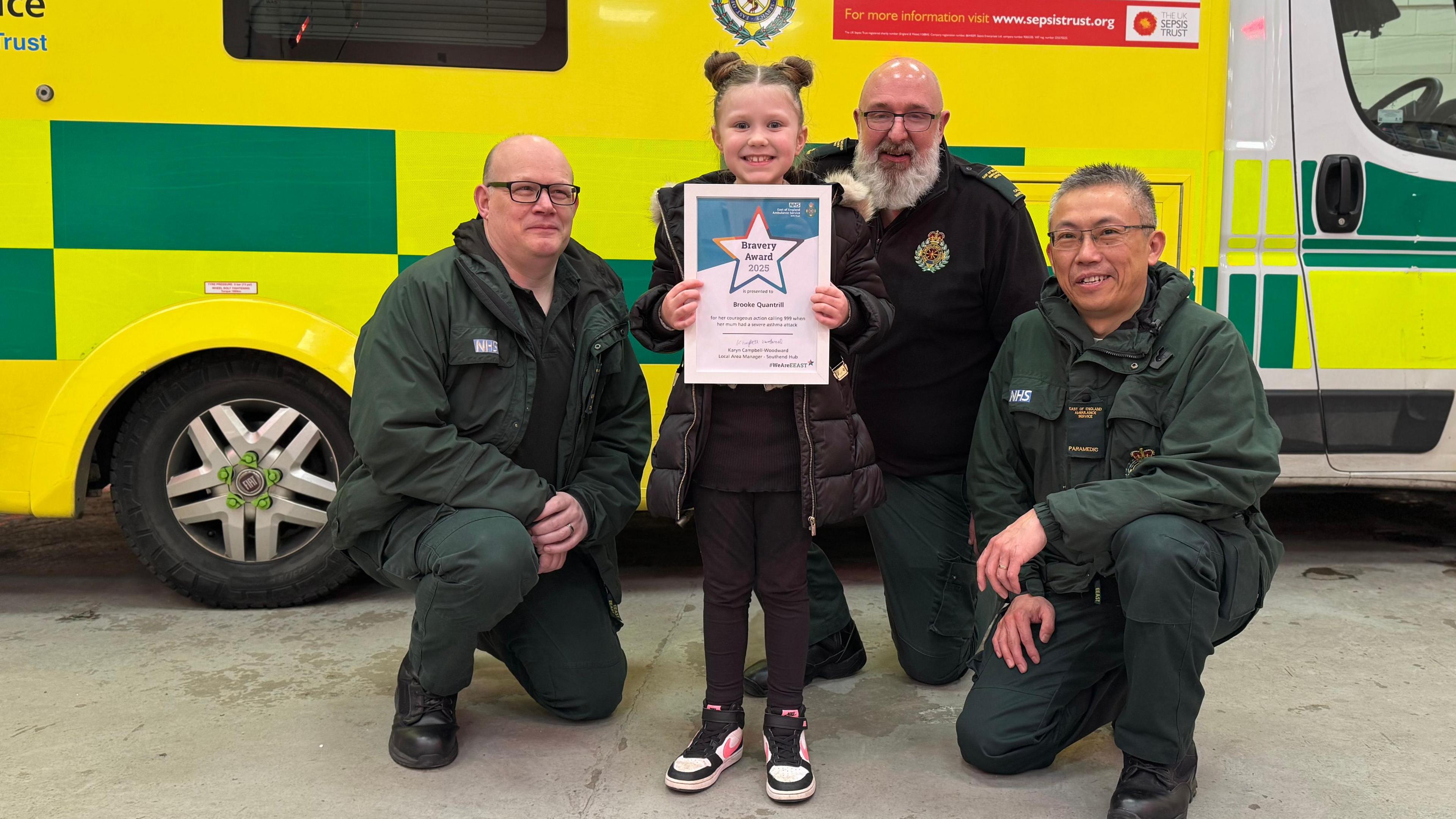 A little girl grinning while holding a framed certificate in front of the call handler with the medics either side of her, behind all of them is a bright yellow and green ambulance