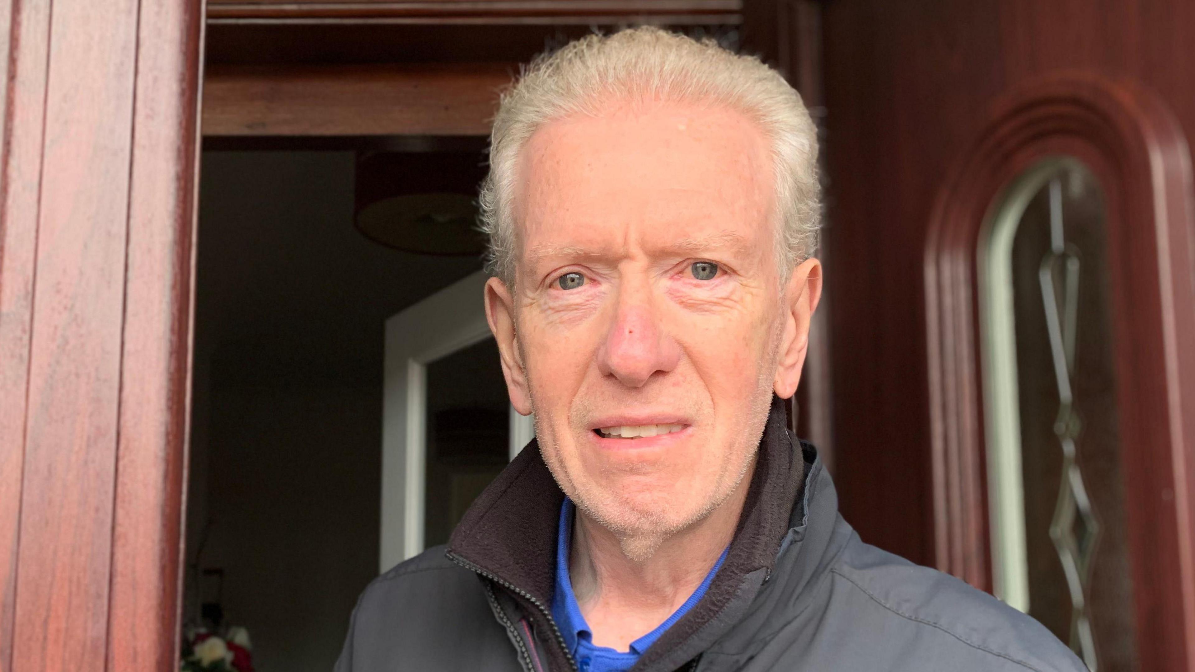 A man with receding grey hair in a black jacket with blue T-shirt underneath stands in a doorway