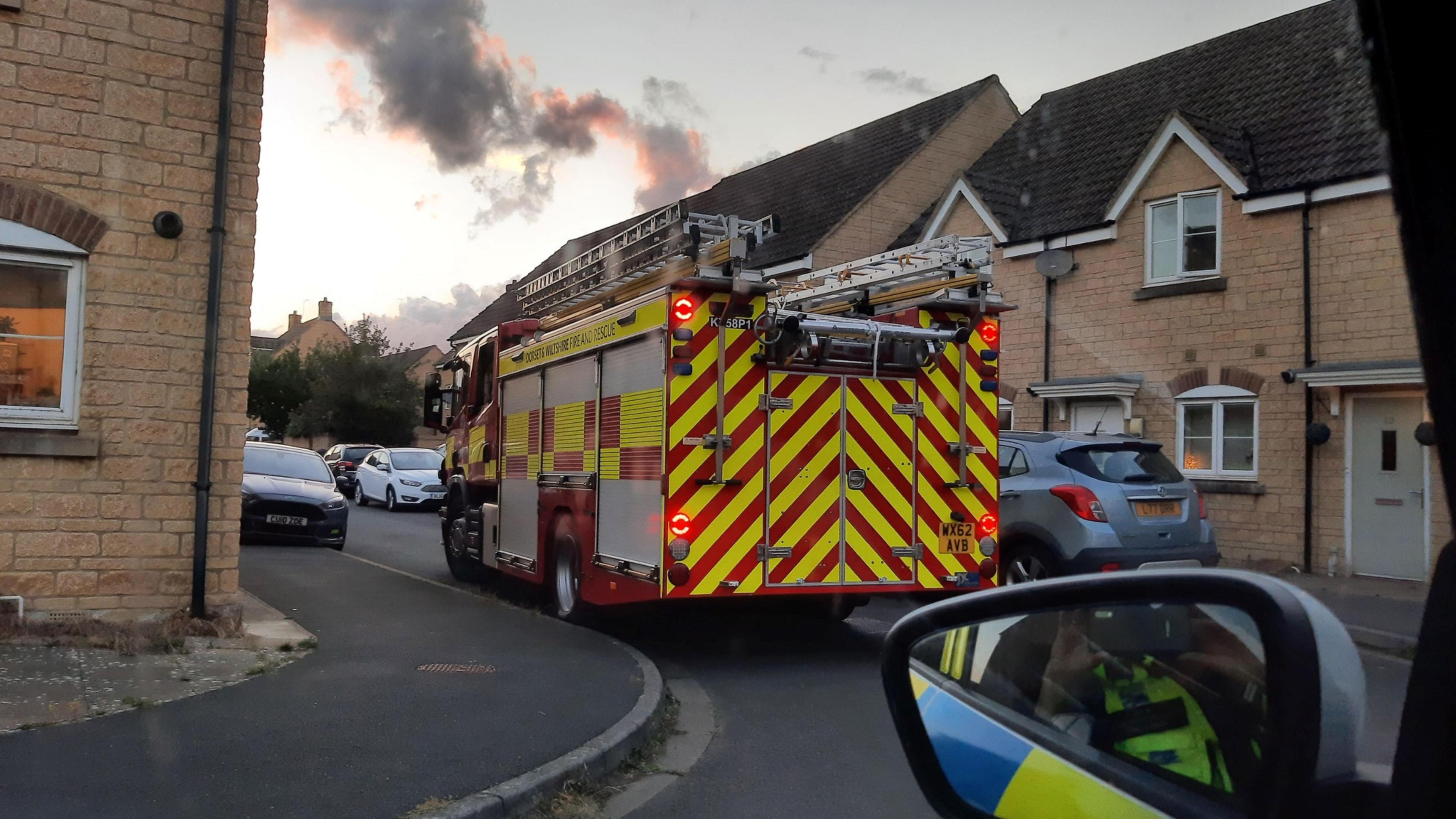 A fire engine navigating through the narrow roads of a housing estate with cars parked on either side of the road. Smoke can be seen in the distance. A wing mirror of a police car is visible on the lower right, with a police officer seen taking the photo.