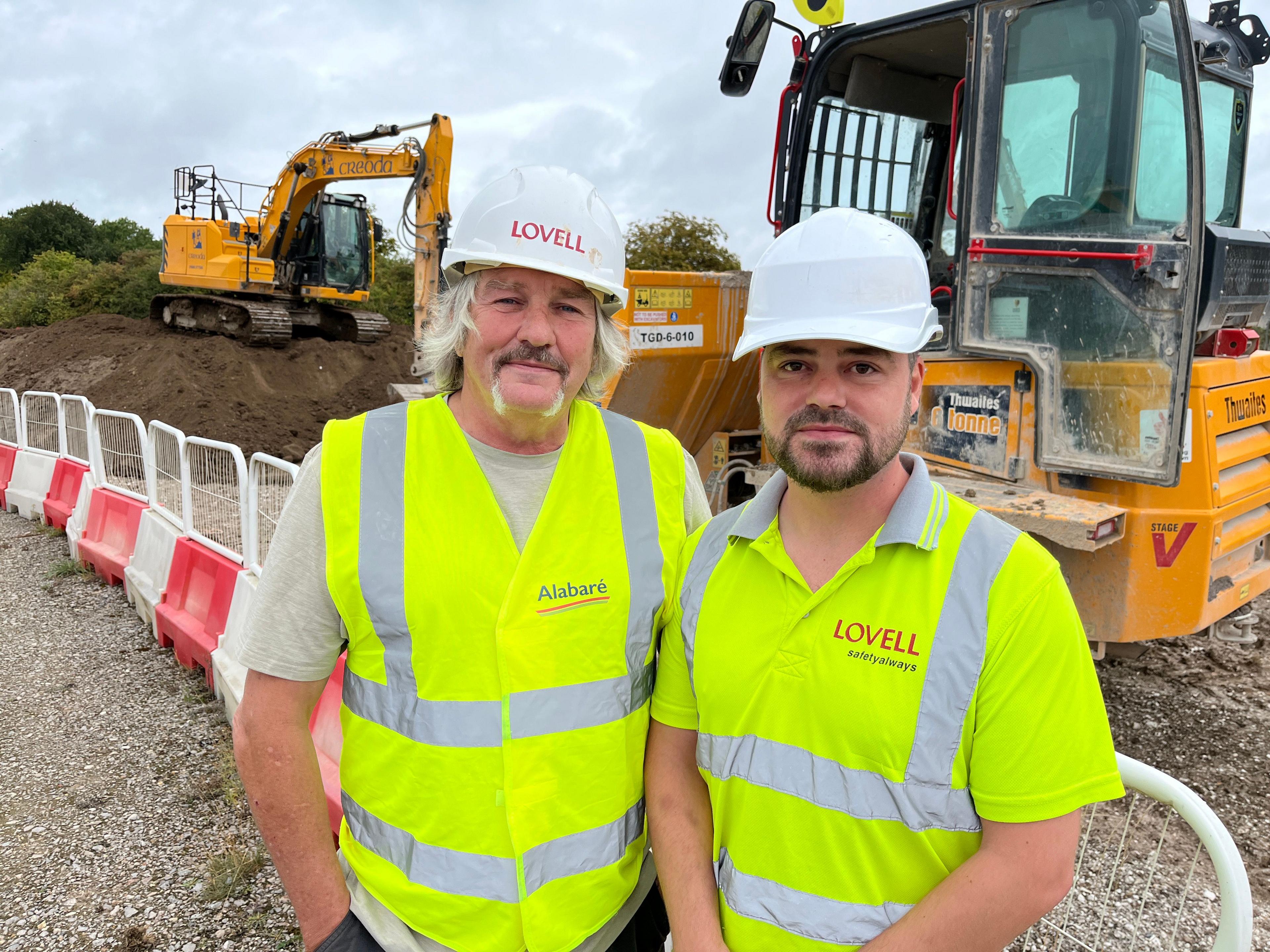 Two construction workers wearing white hardhats and yellow high visibility tops standing in front of two bulldozers
