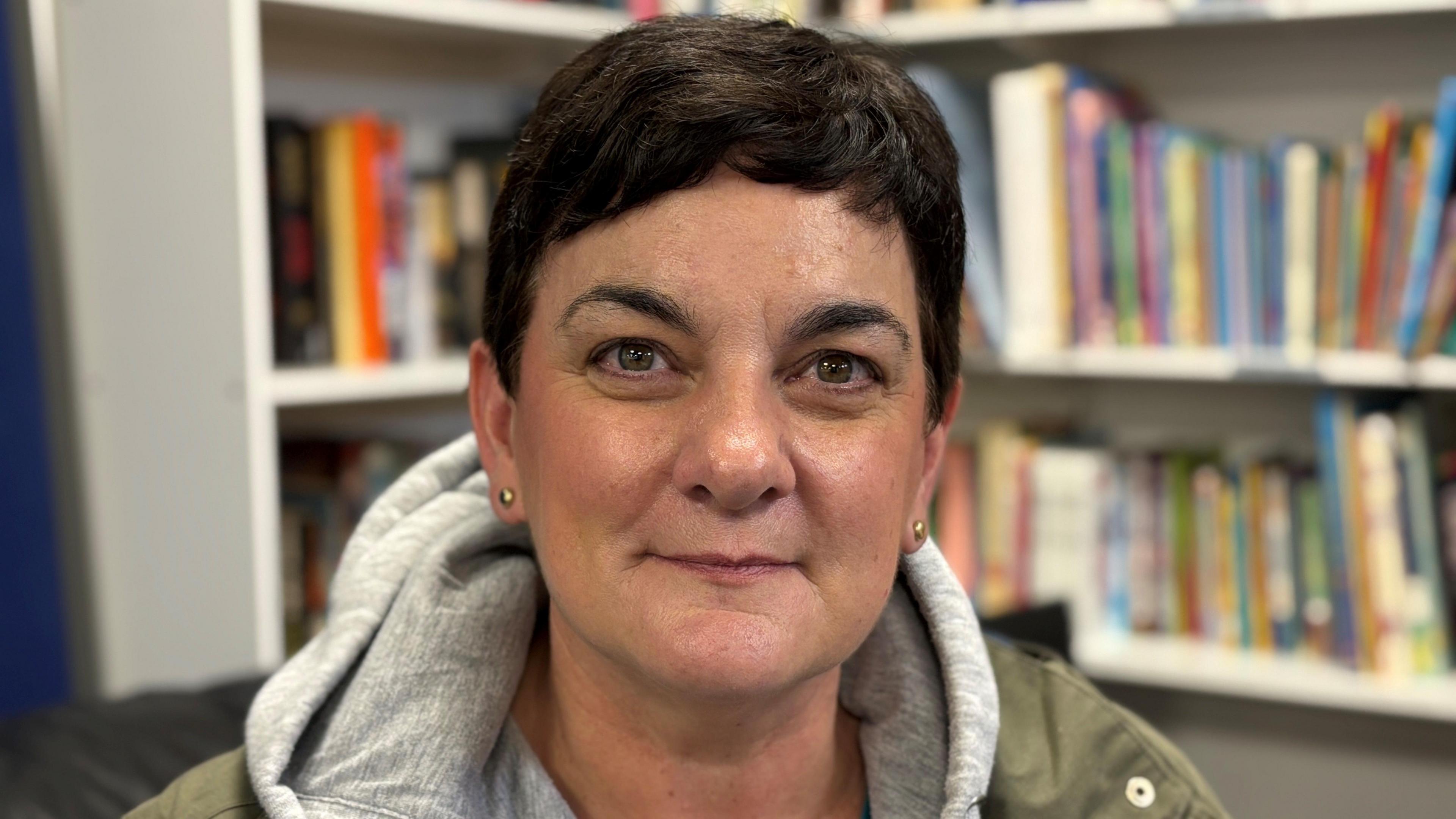A woman with short black hair wearing a grey hoody and green jacket. There are bookshelves with books behind her.