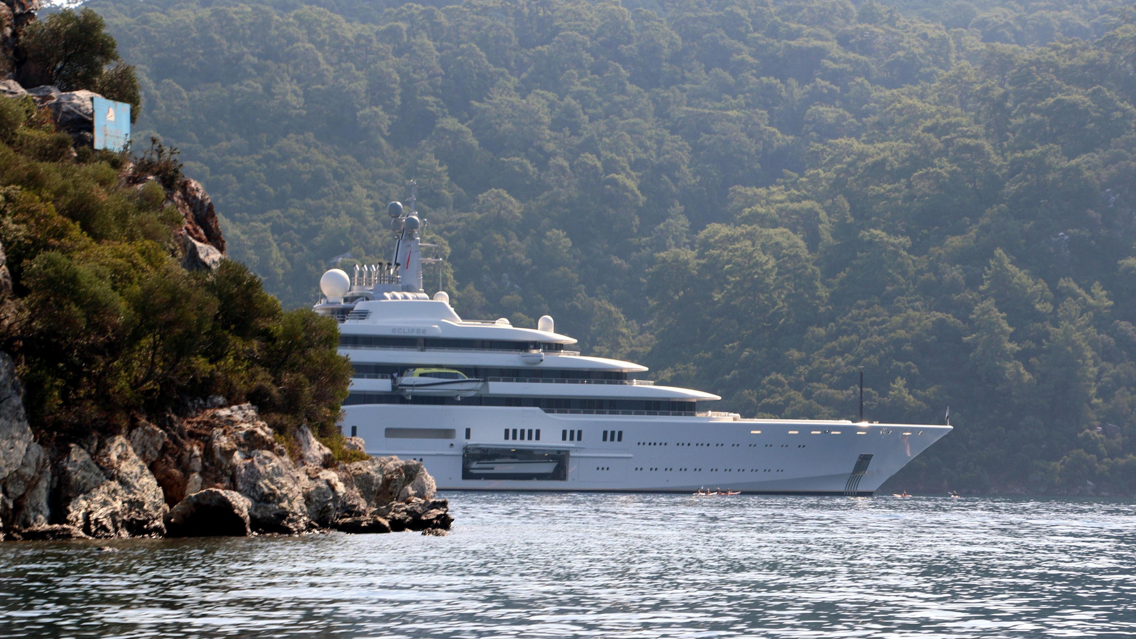 The super-yacht Eclipse, emerging from behind a rocky outcrop in the Mediterranean near Turkey, with a wooded hillside behind it. A door on the side of the yacht is open to reveal a smaller vessel inside