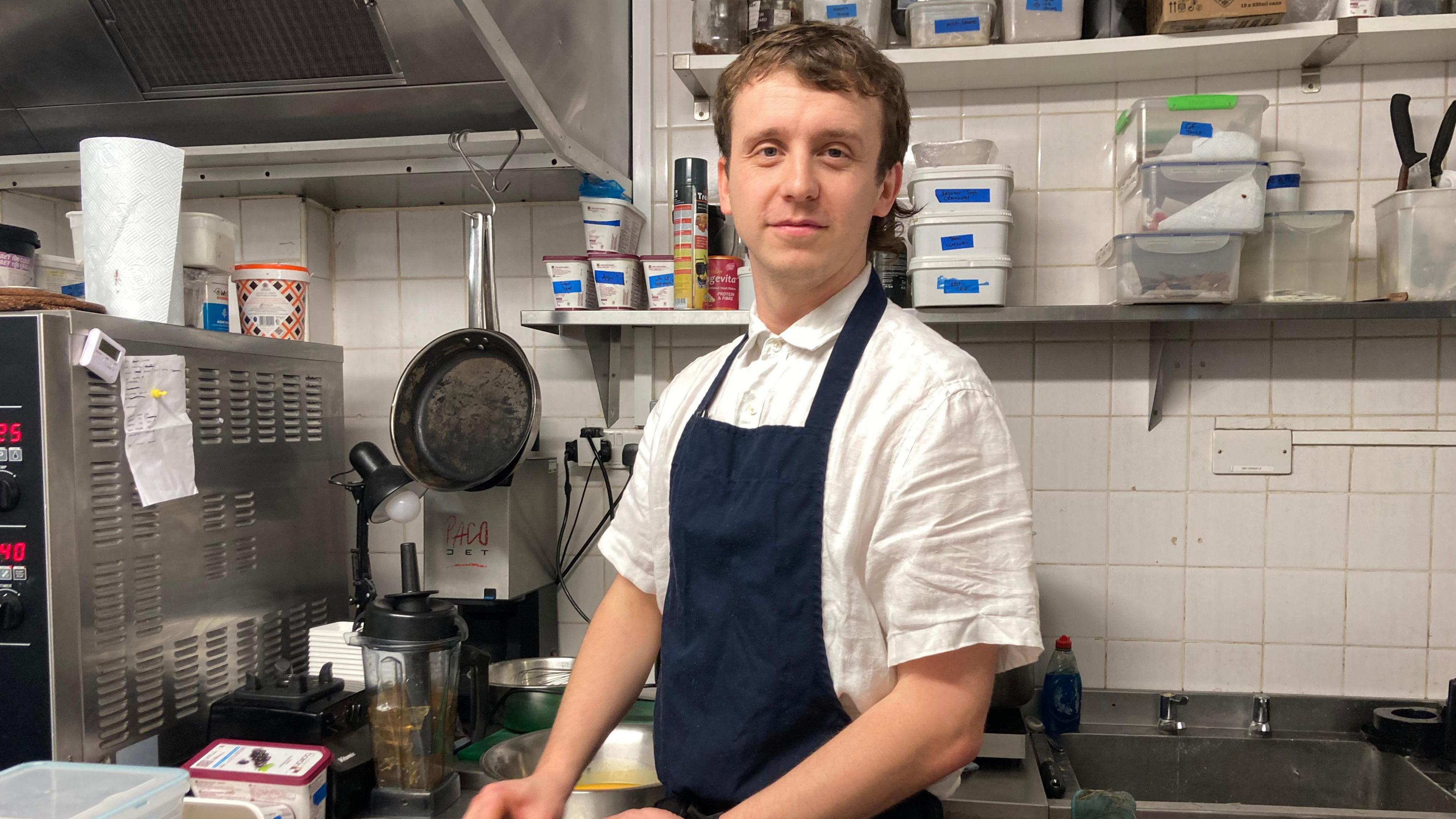 Ben Willcocks - A chef is wearing a navy apron and looking at the camera. He is standing in a kitchen and surrounded by equipment.