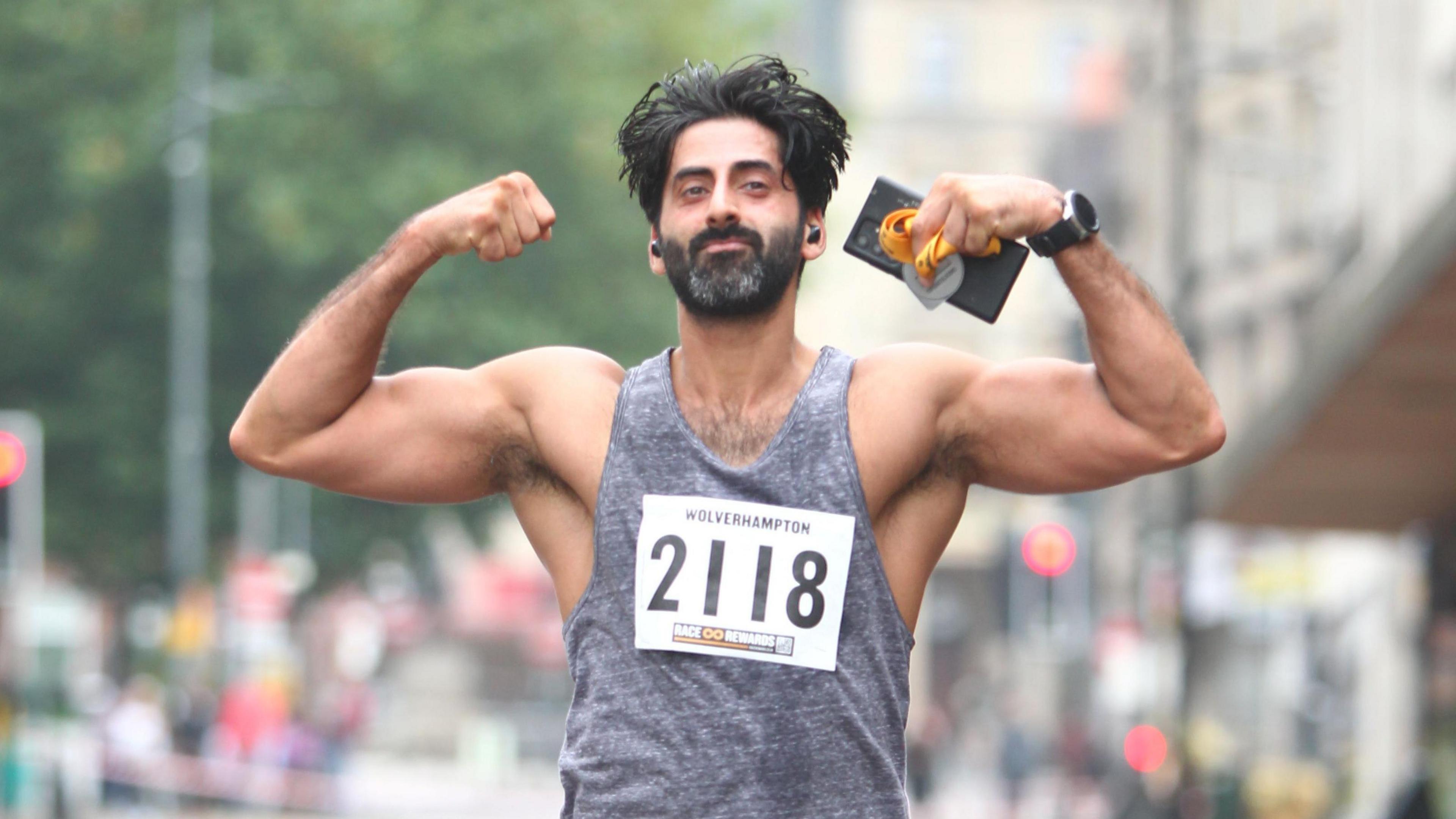 A man in a grey top raises his arms, he is holding a phone and medal in his hand