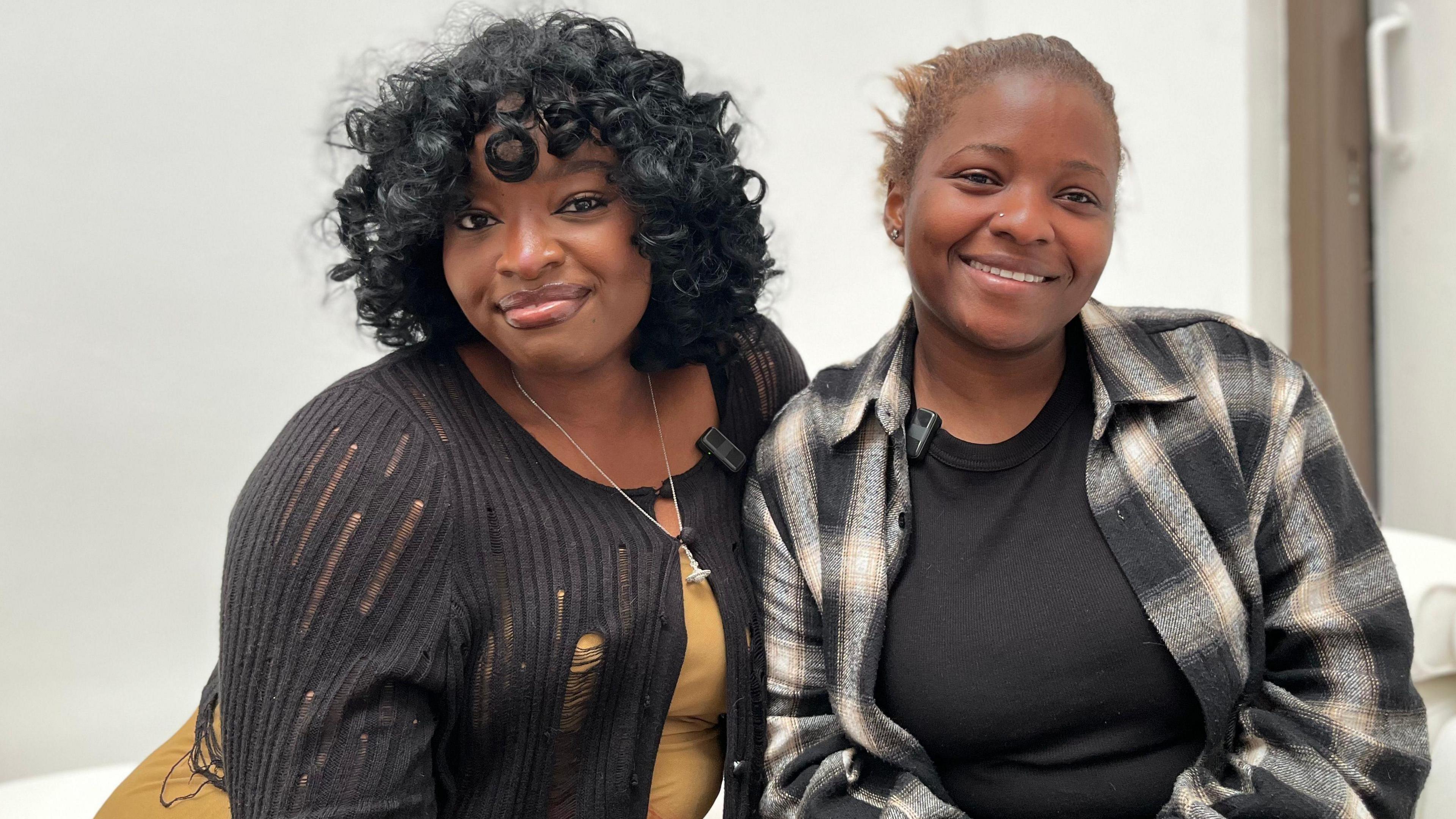 Two women are sitting next to each other and smiling. The woman on the left is wearing a black cardigan, a yellow top and has black curly hair. The woman on the right is wearing a plaid jacket and a black top.