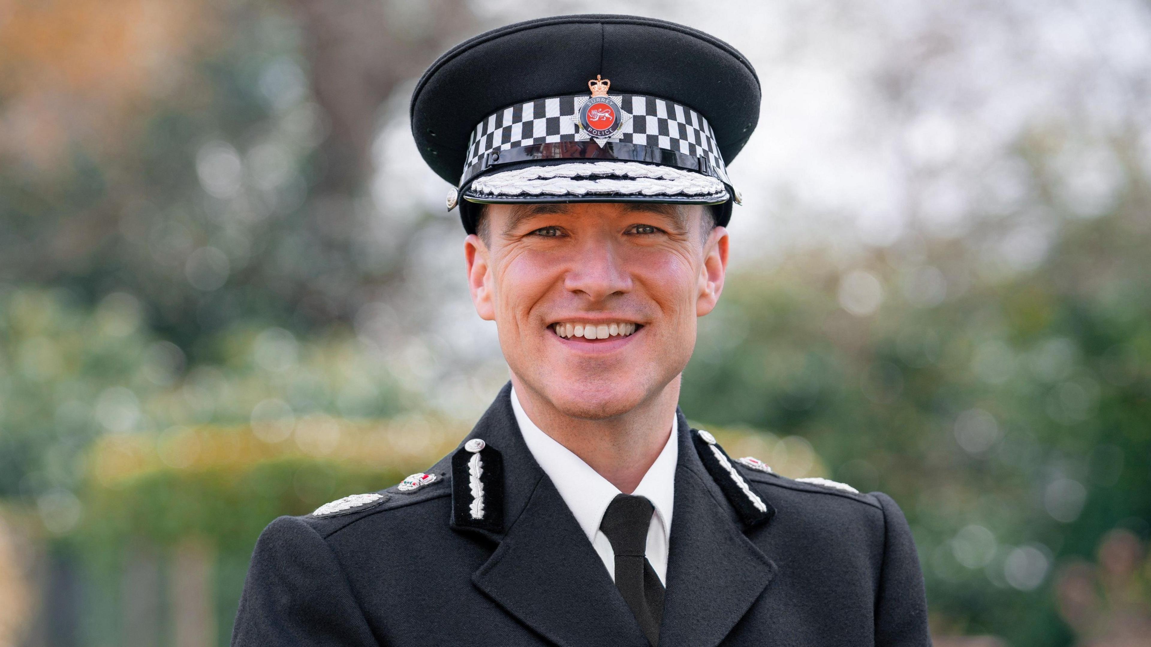 Surrey Chief Constable Tim de Meyer smiles into the camera, wearing his full dress tunic, white shirt, black tie and cap.