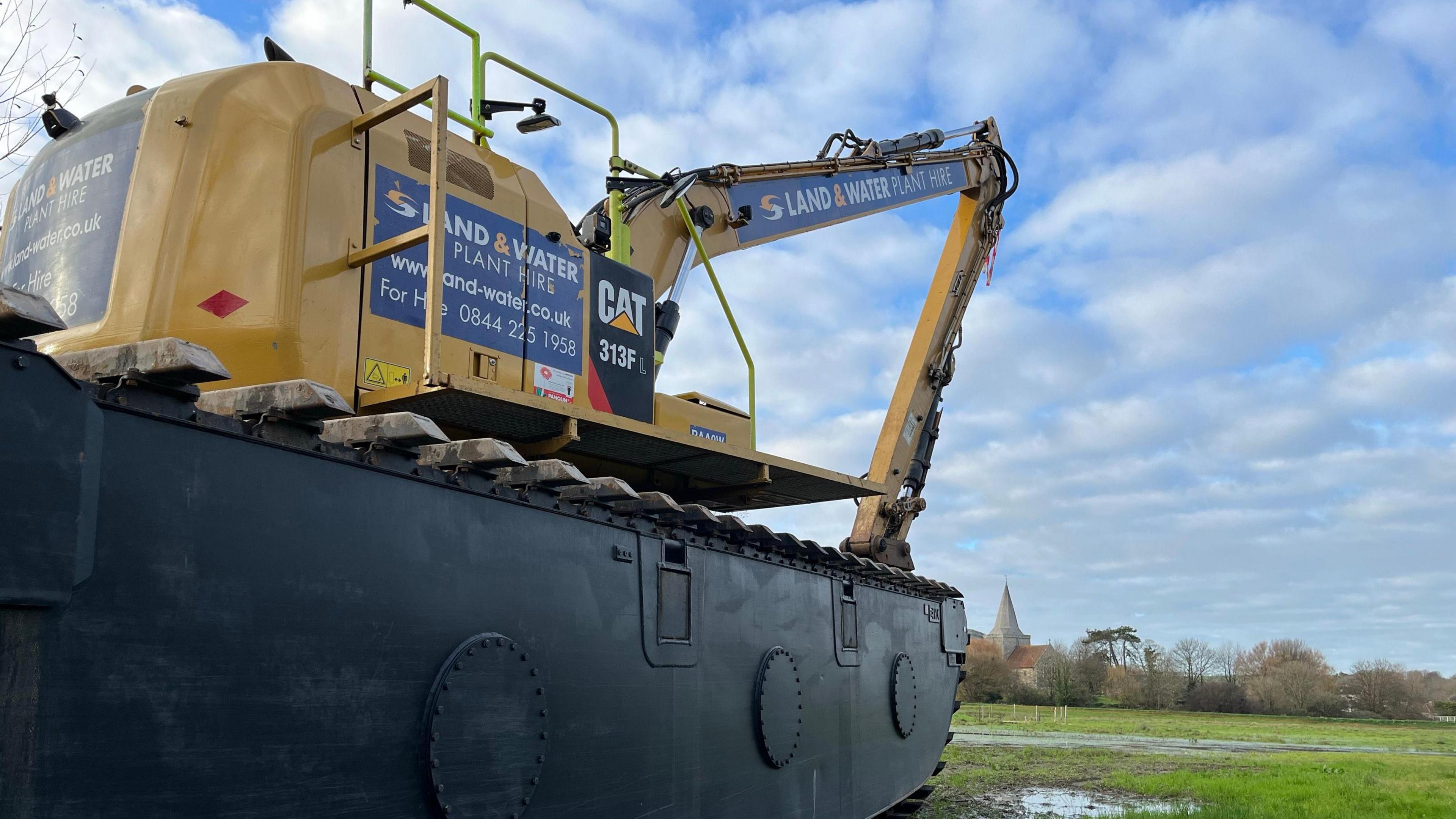 A yellow amphibious digger which can float on water in a field near Alfriston Church which is in the background