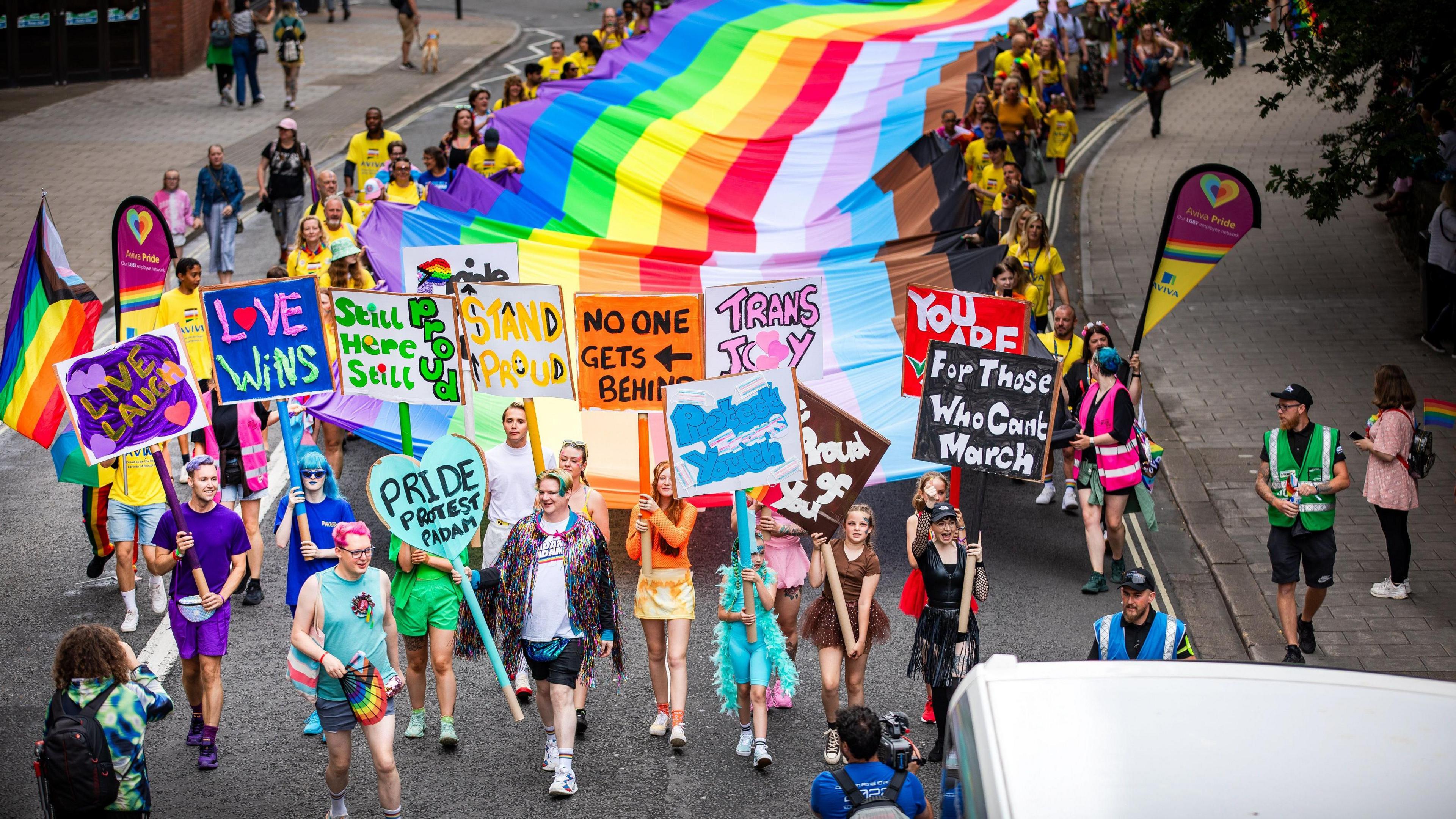 People holding banners in support of LGBT+ people, with a long rainbow banner stretching behind them. 