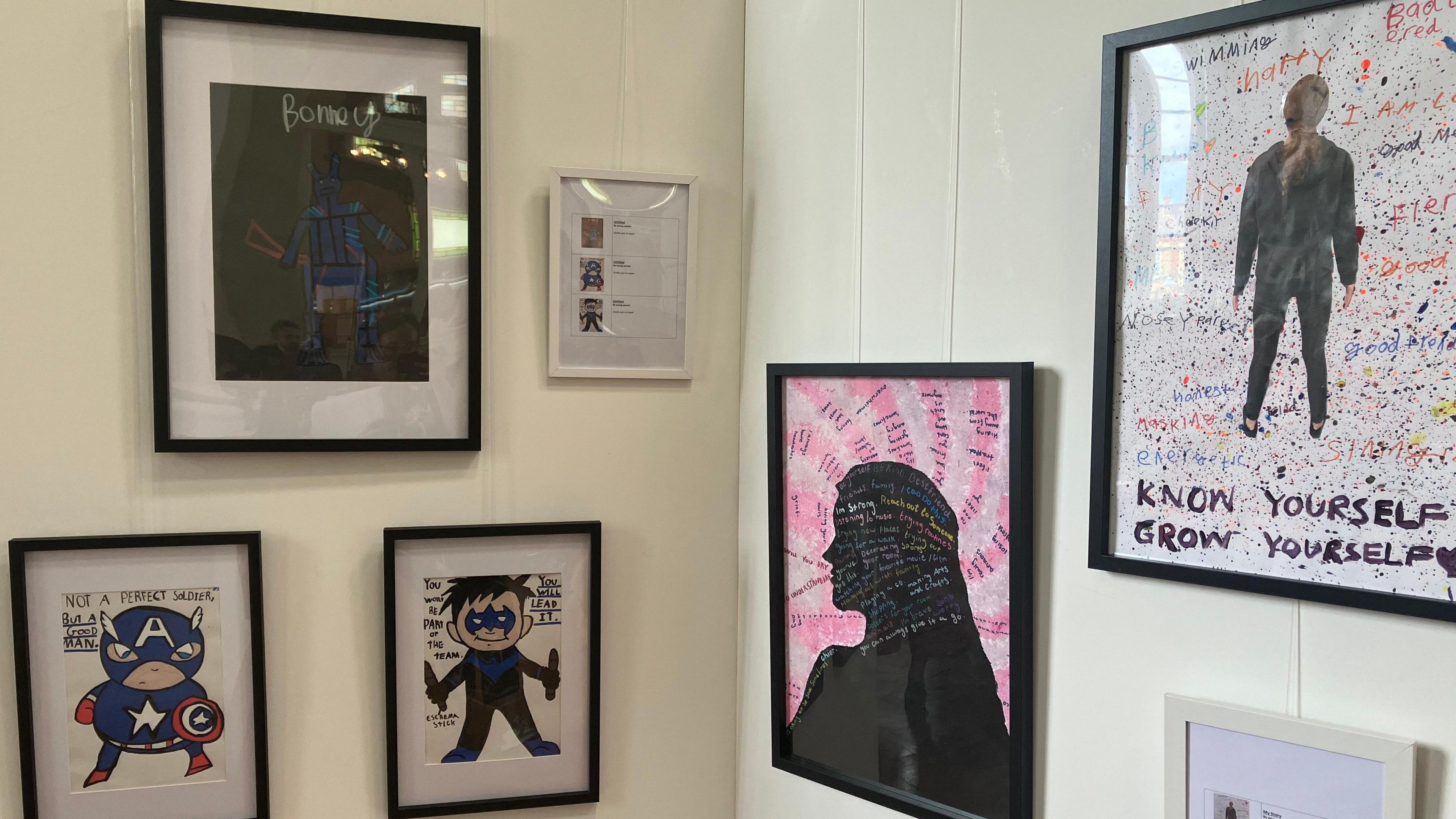 A display of five pieces of art on a white wall. Two of them are superheroes and two others are silhouettes of people on a background of words.