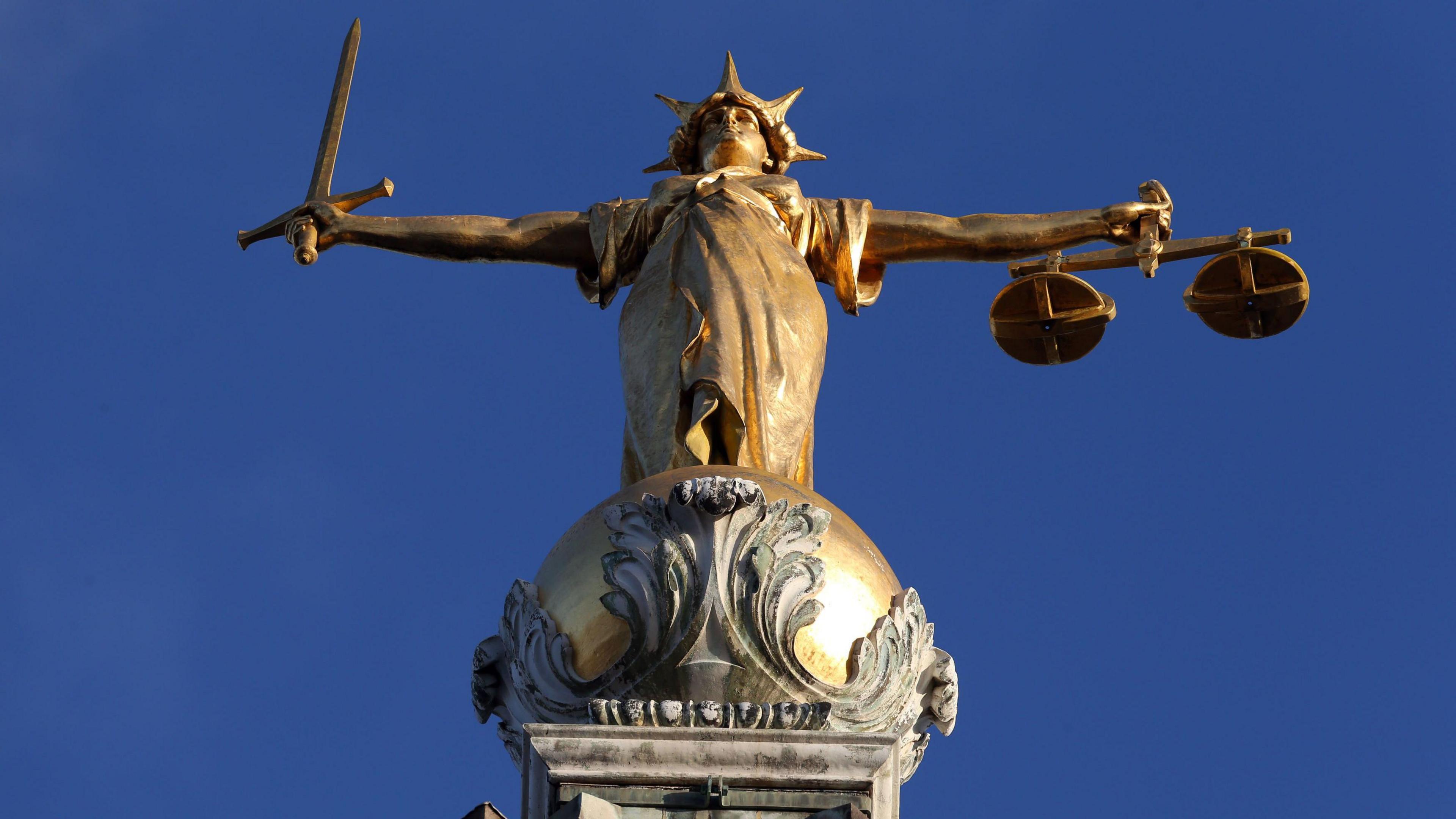 A golden statue of a person wearing a crown, holding a sword and a set of scales on top of a golden ball, which sits on a plinth.