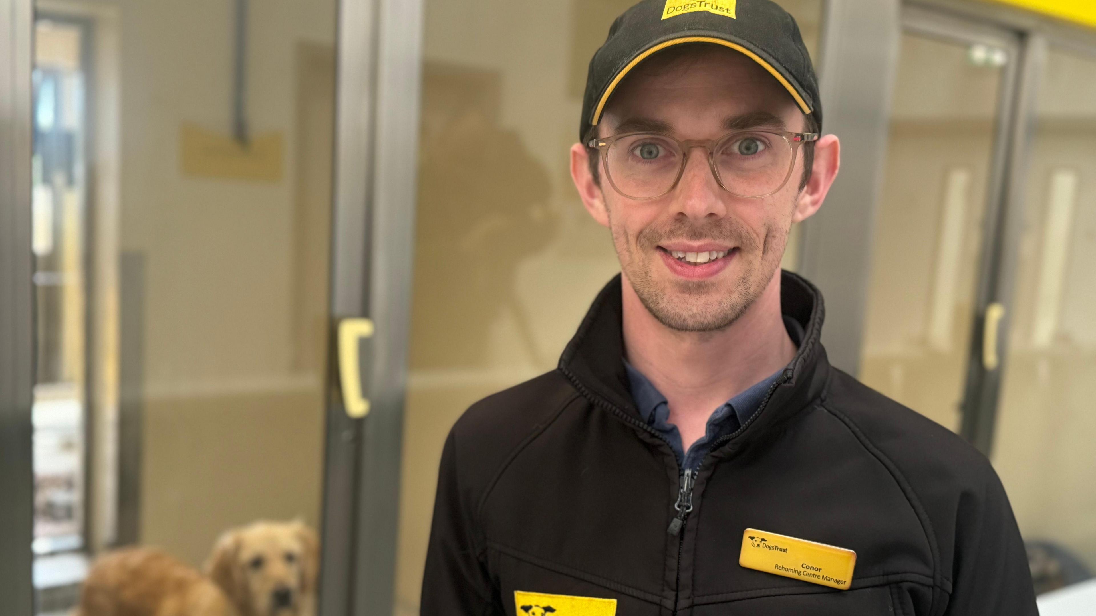 Conor smiling at the camera. He is wearing glasses and a baseball cap. His jacket is black and he has a yellow name badge. There is a dog in the background.