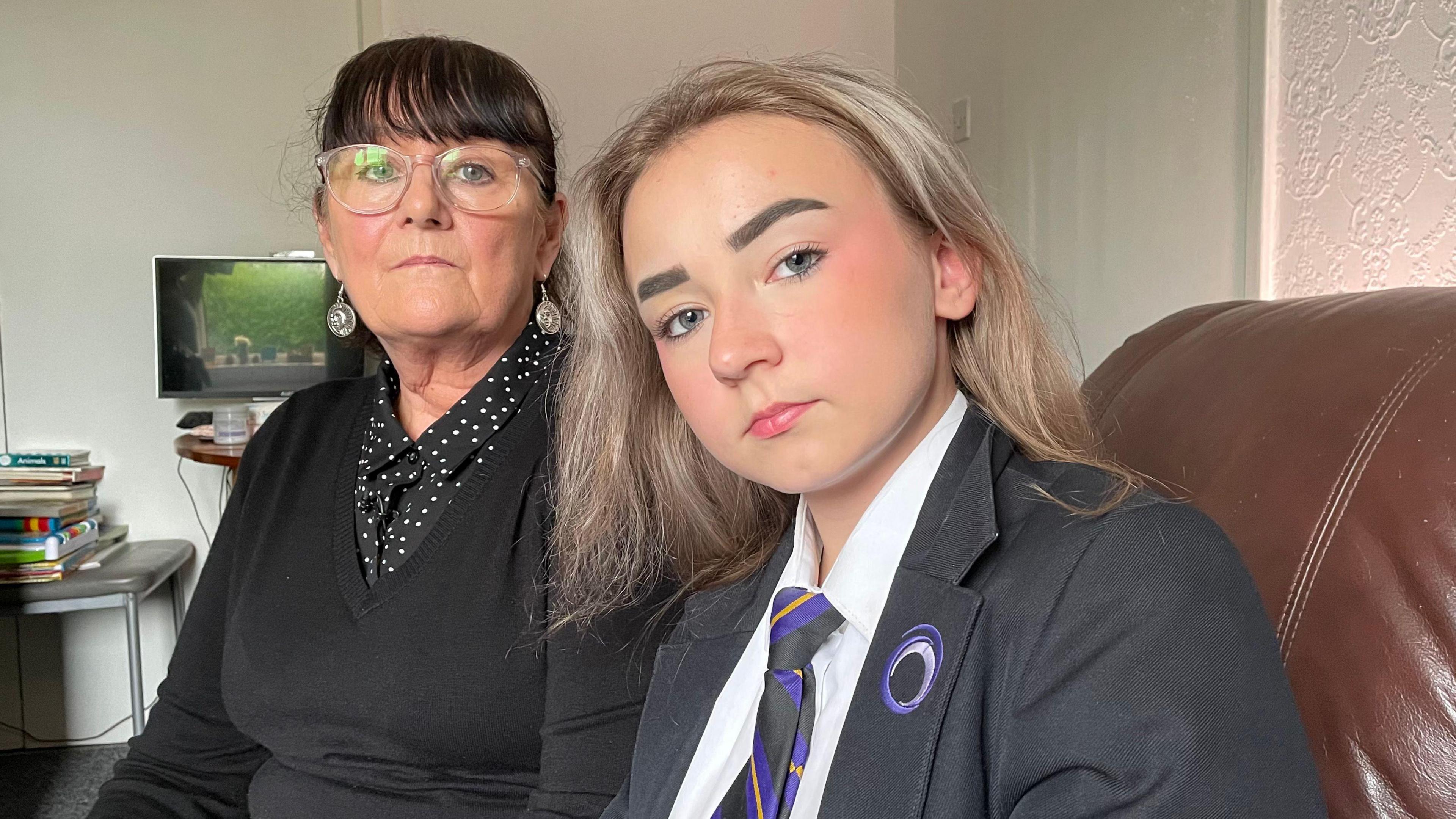 A woman with dark hair and glasses sits next to her granddaughter on a sofa at home. The granddaughter wears her school uniform, including a blazer and tie.