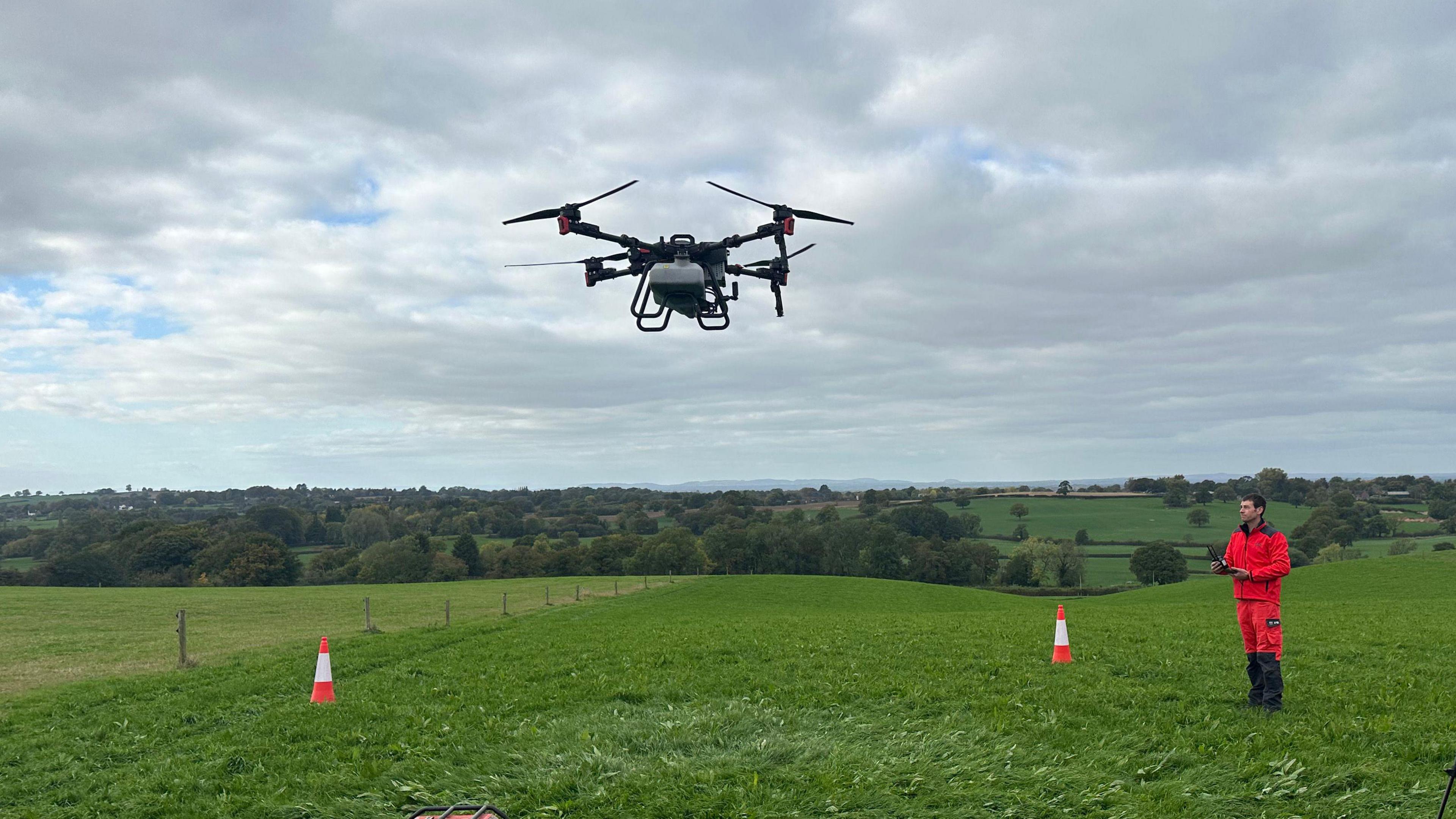 A man wearing bright red overalls and wellies, stands in a massive field on a hillside while holding controls. A large black drone is hovering in the air. Two traffic cones have been placed on the field. On the horizon are fields and trees stretching into the distance.