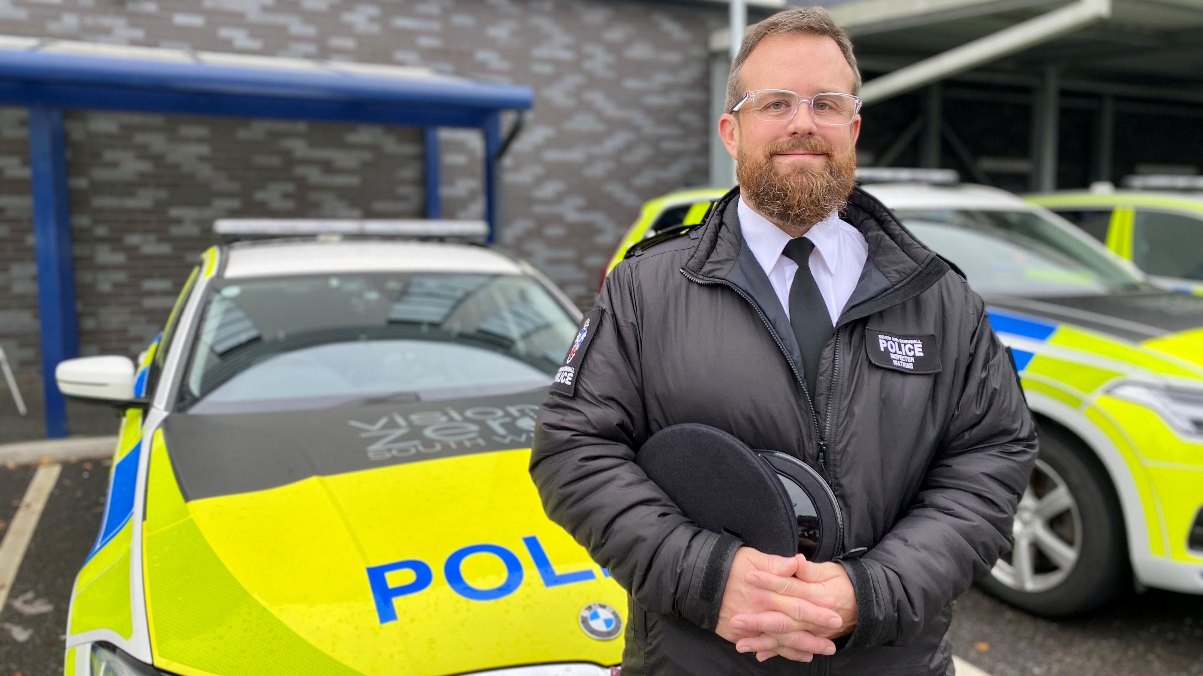 Patrol Insp Wes Watkins standing in front of two police cars. He is a beard and is wearing glasses, a tie and is holding his hands together.