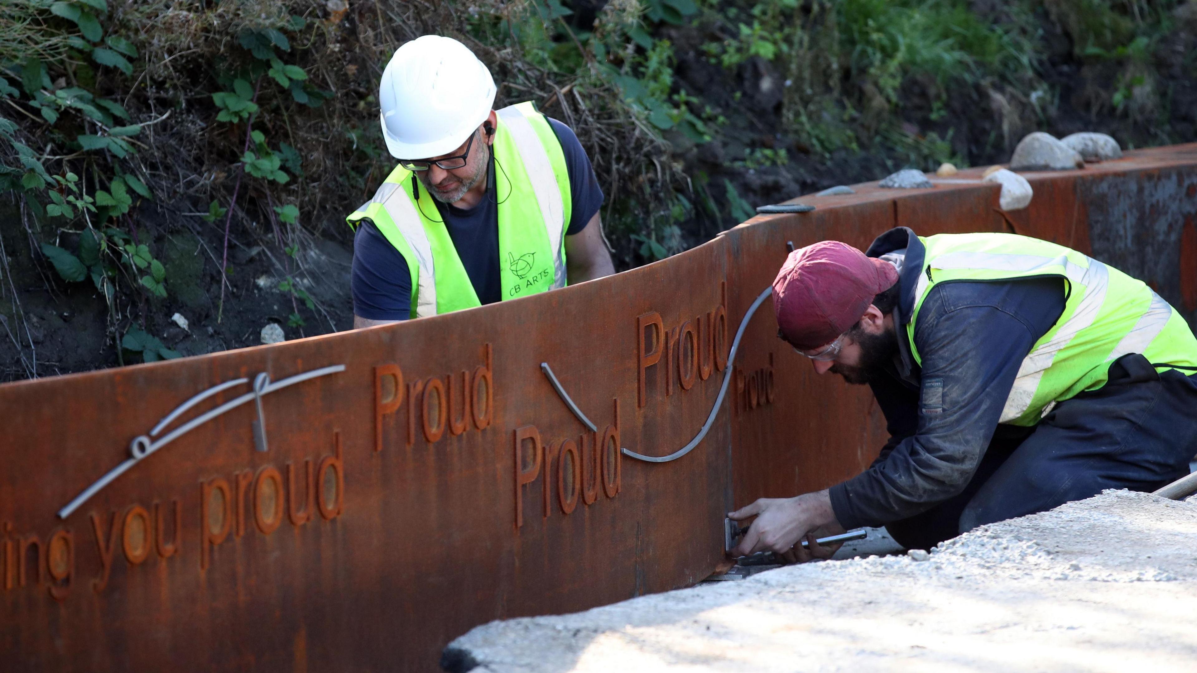 Two workman installing the artwork in Roker Park. The word "proud" is shown several times on the rusted-looking metal.