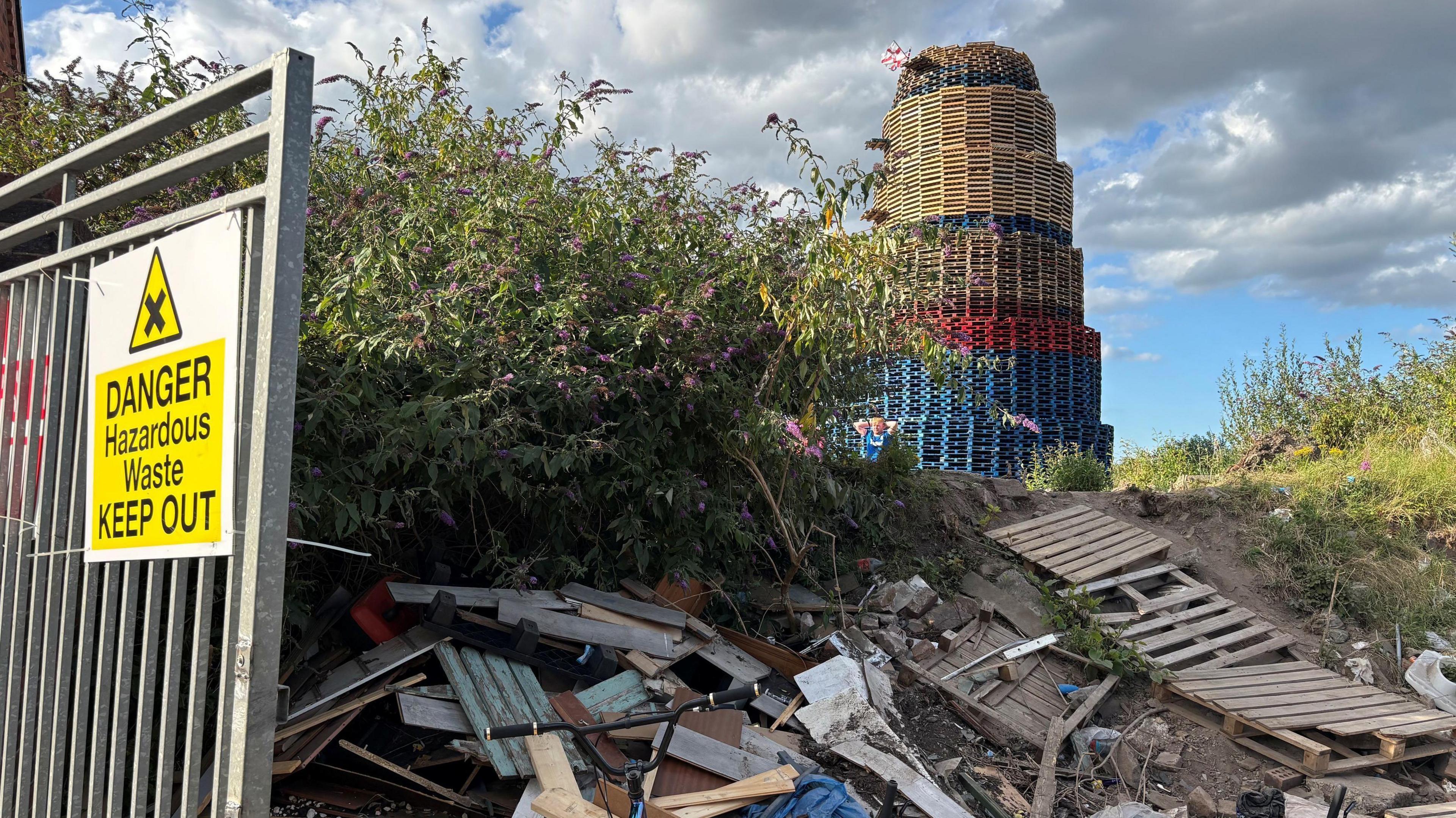A rubbish pile lies behind a mental gate which has a sign that says 'Danger Hazardous Waste Keep Out' on it. In the background there is a towering bonfire made out of wooden pallets.
