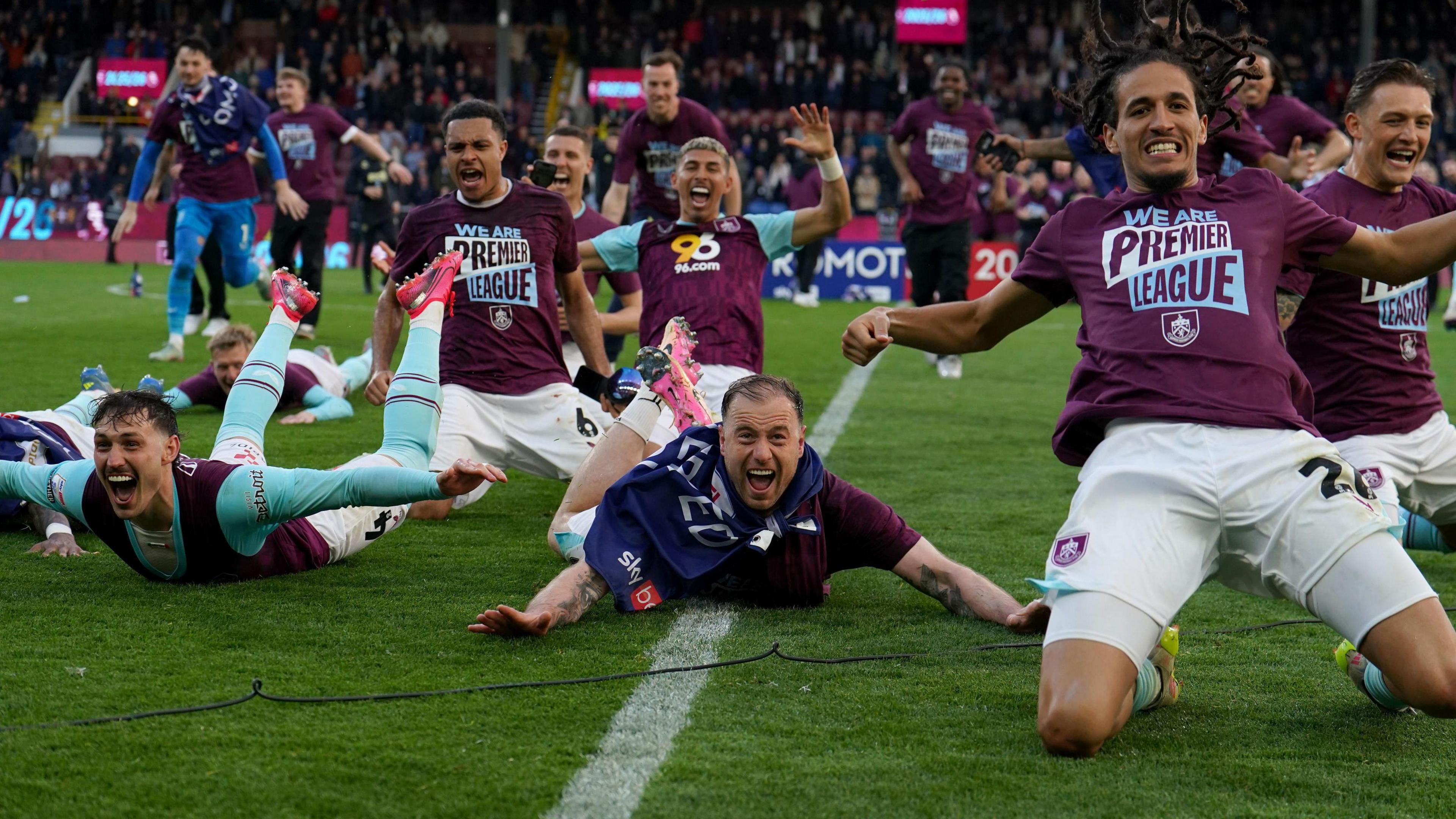 Burnley players slide across the Turf Moor pitch in celebration wearing t-shirts with the slogan 'We Are Premier League'