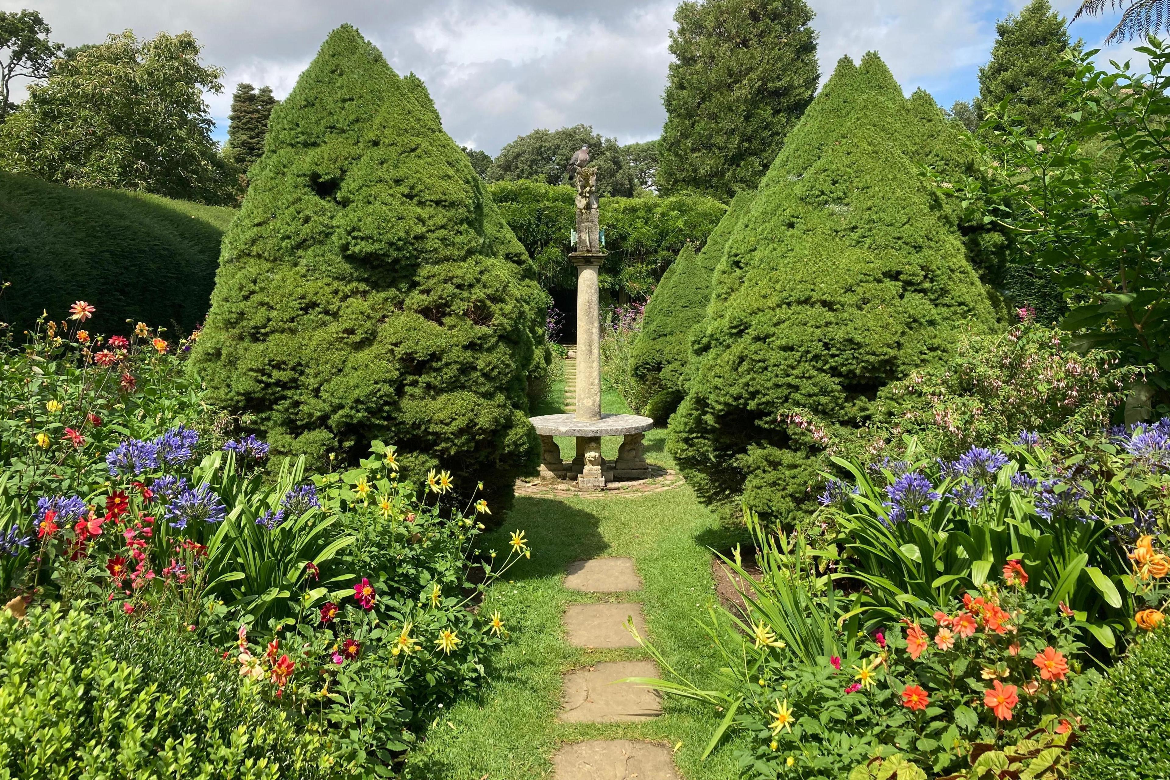 A walled garden view of a stone path between beds of colourful flowers leading to a stone bench with a tall plinth rising from it's centre. A wood pigeon sits on it's top. Rows of conical shaped spruce trees stand either side of the bench