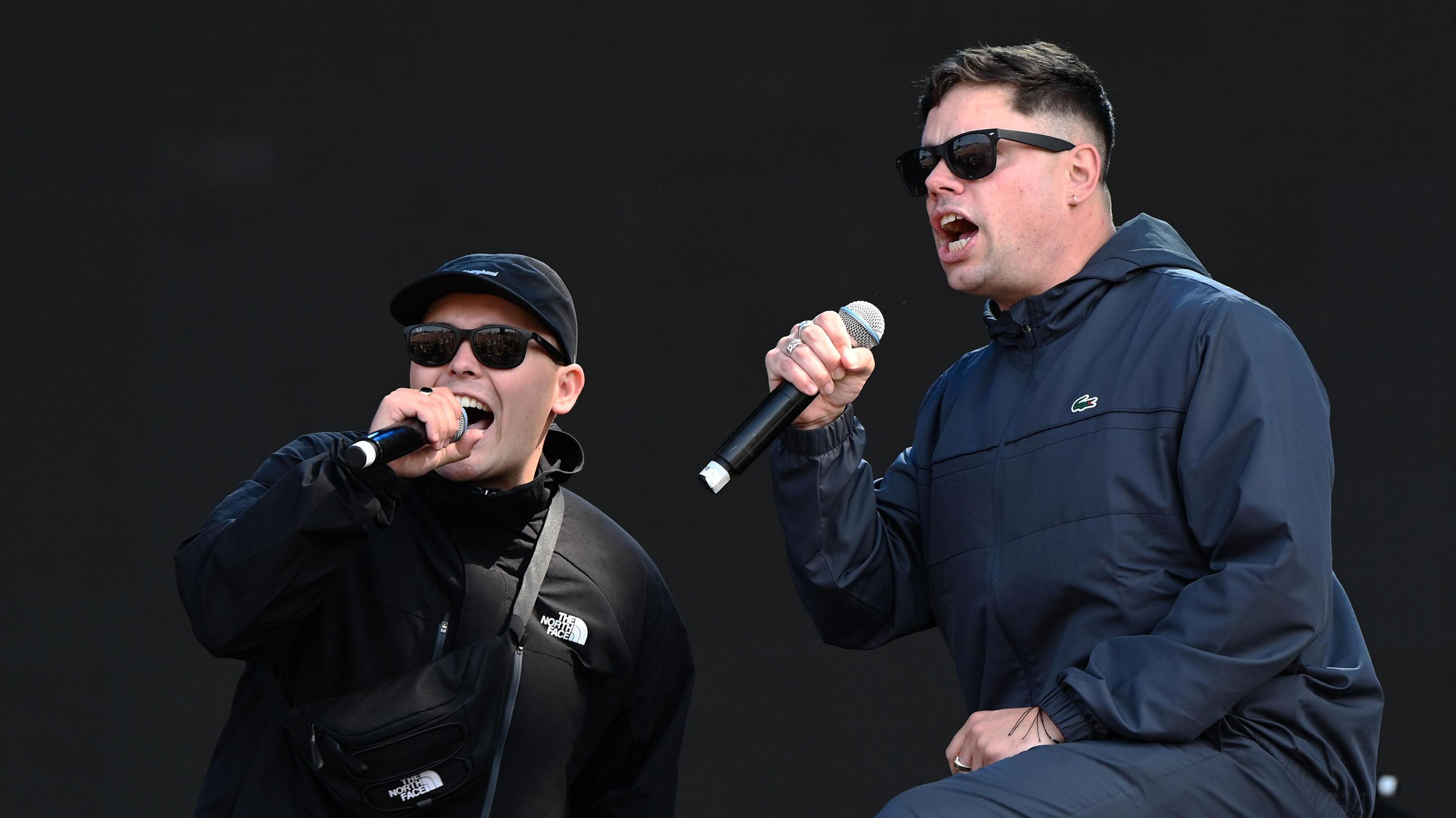 Kneecap performing at Reading Festival in August 2024.
Two members of kneecap are holding microphones. One is wearing a black jacket, black baseball hat and sunglasses.
The man on the righ is wearing a navy top and trousers and a pair of sunglasses.