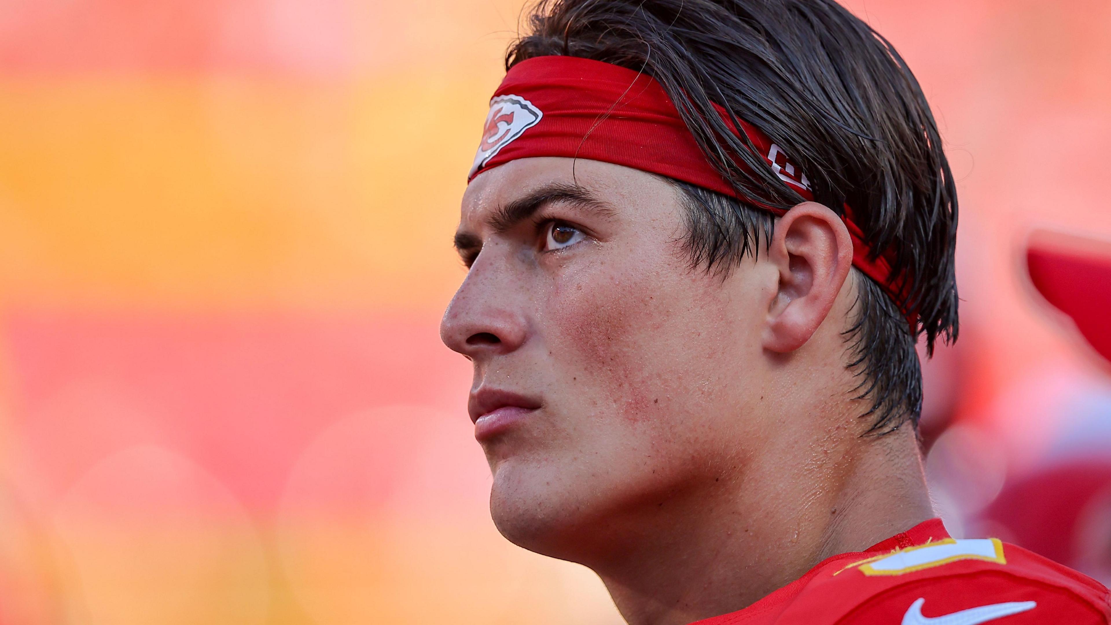 Louis Rees-Zammit at an NFL game, wearing a red Kansas City Chiefs headband to push back his dark brown hair. He wears a red football shirt and is flushed and sweaty with a serious expression. 