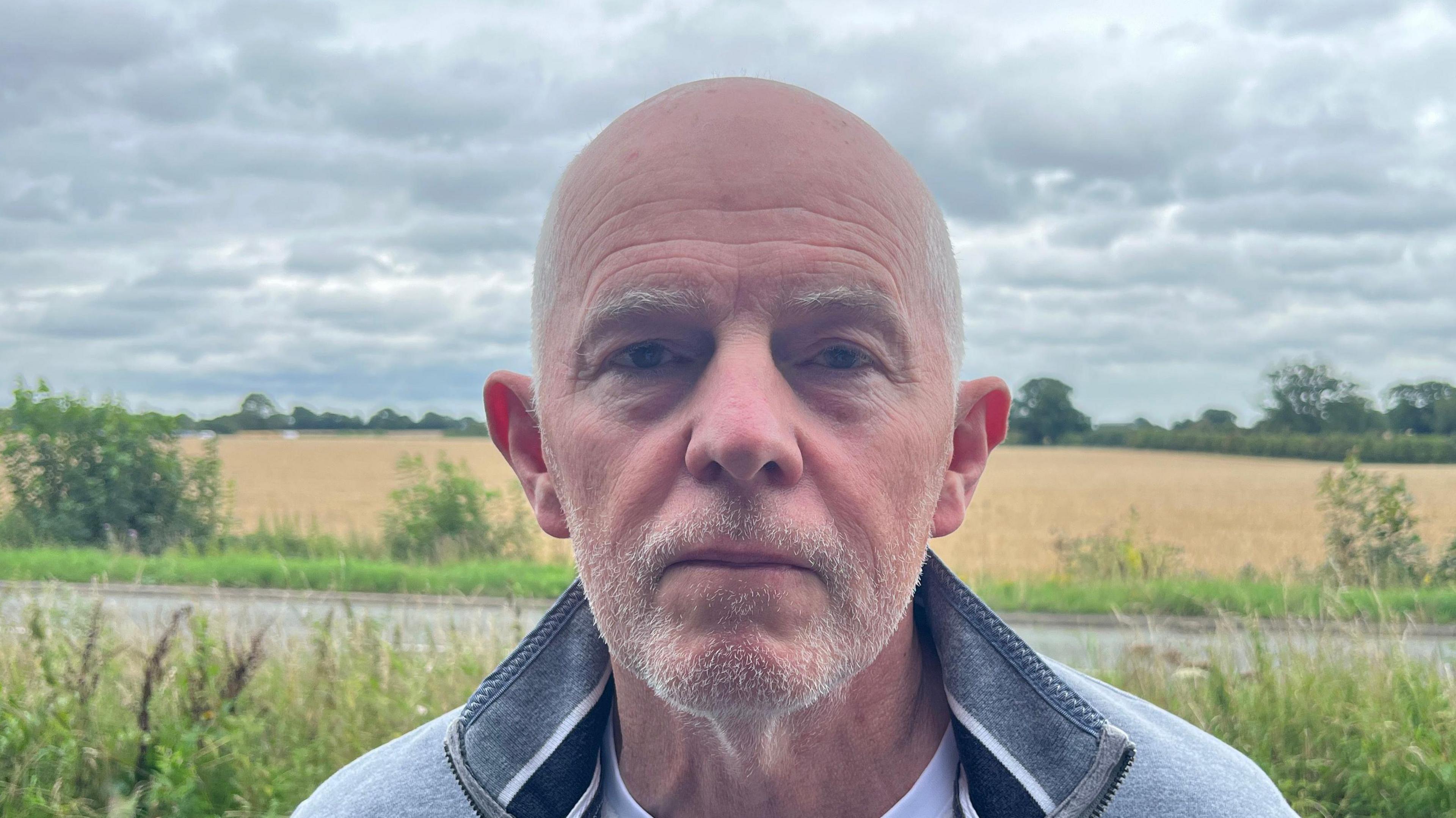 A head and shoulders close-up photo of Malcolm MacDonald from Stop The Quarry. He is wearing a grey jumper and standing in front of a countryside landscape.
