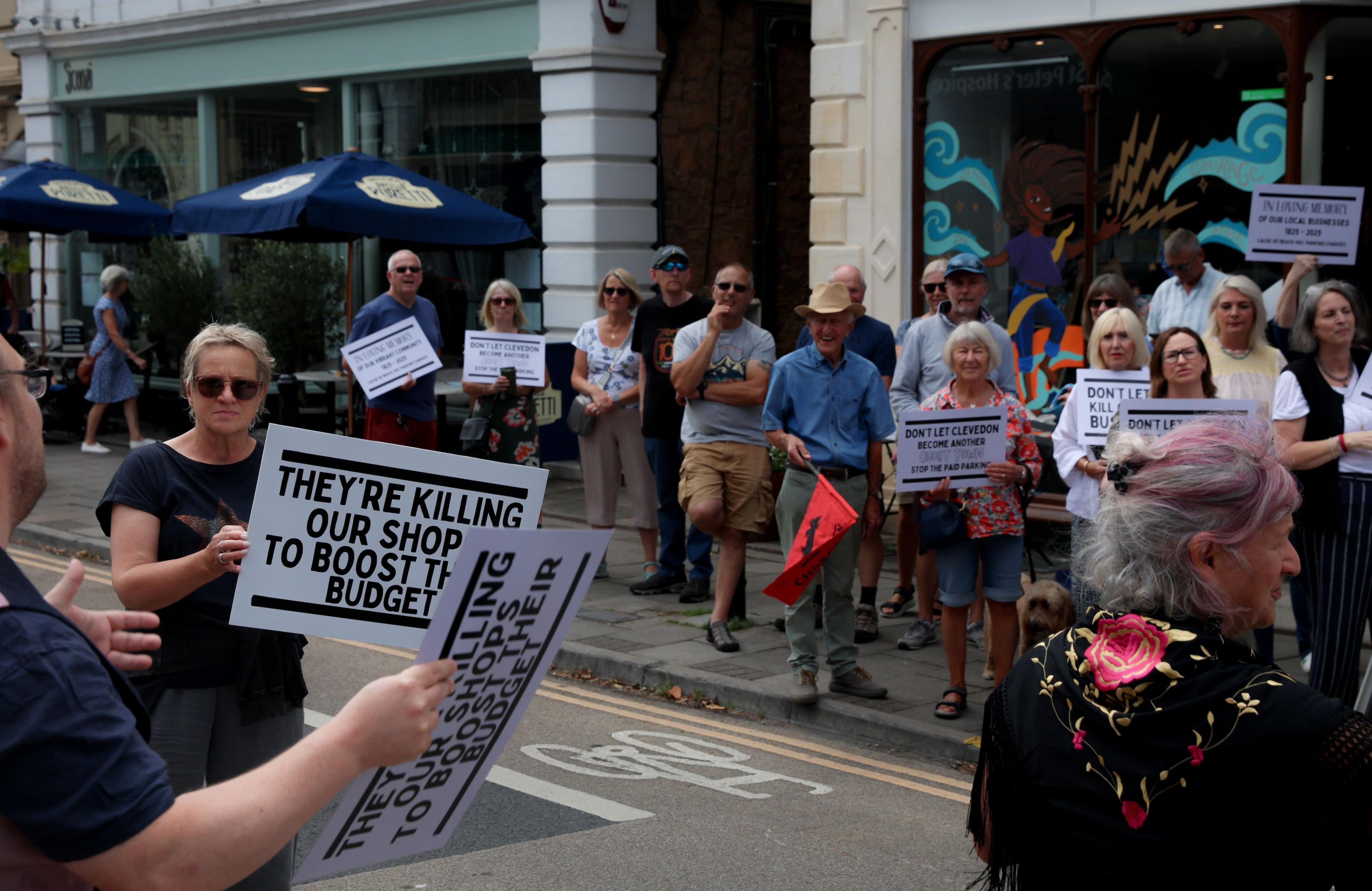 Protesters gather in a street. Some are holding placards that say 'they're killing our shop to boost their budget'.