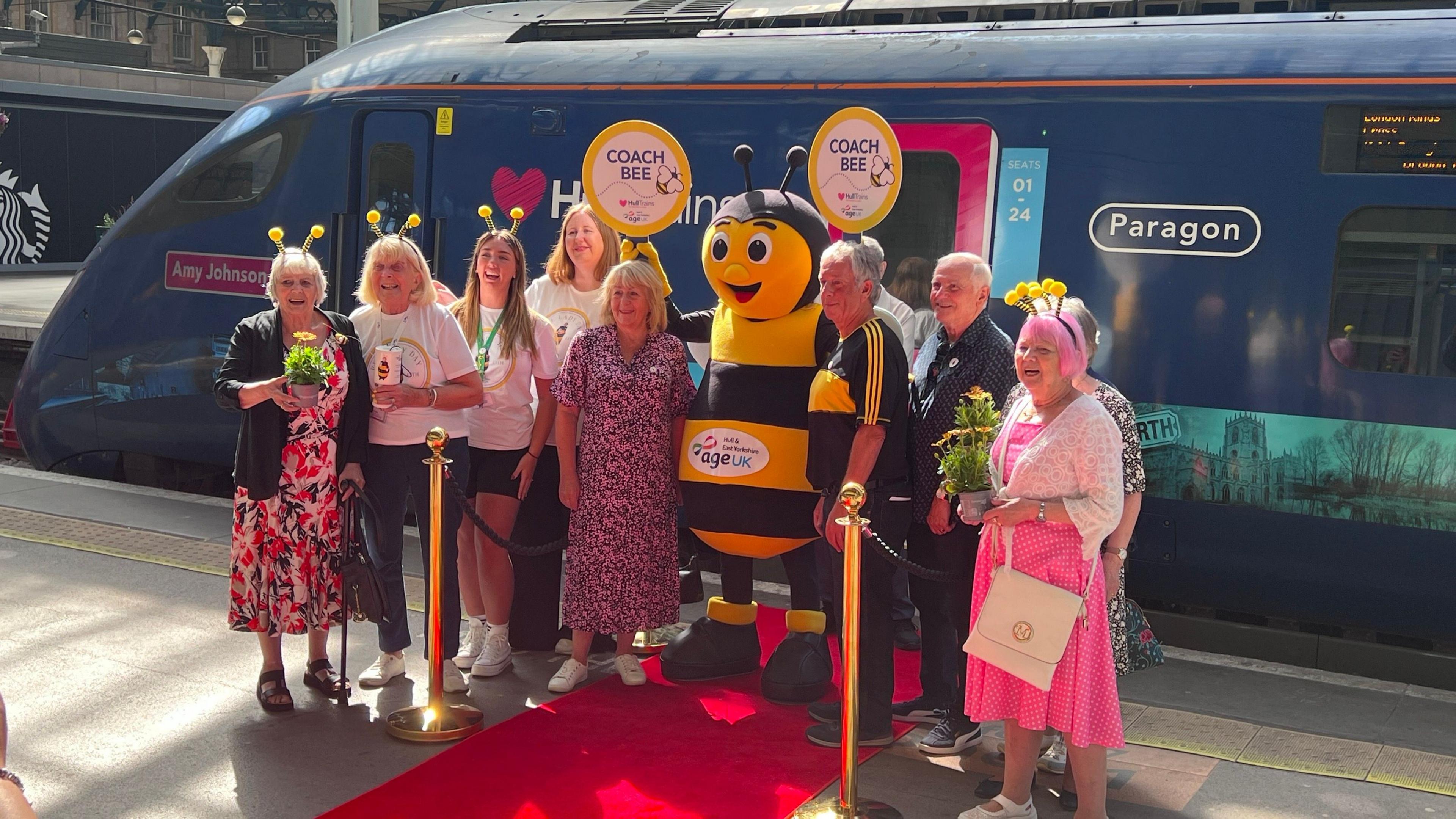 A group of people, many wearing colourful outfits and bee headbands, and someone dressed as a bee, standing in front of train on a railway platform.