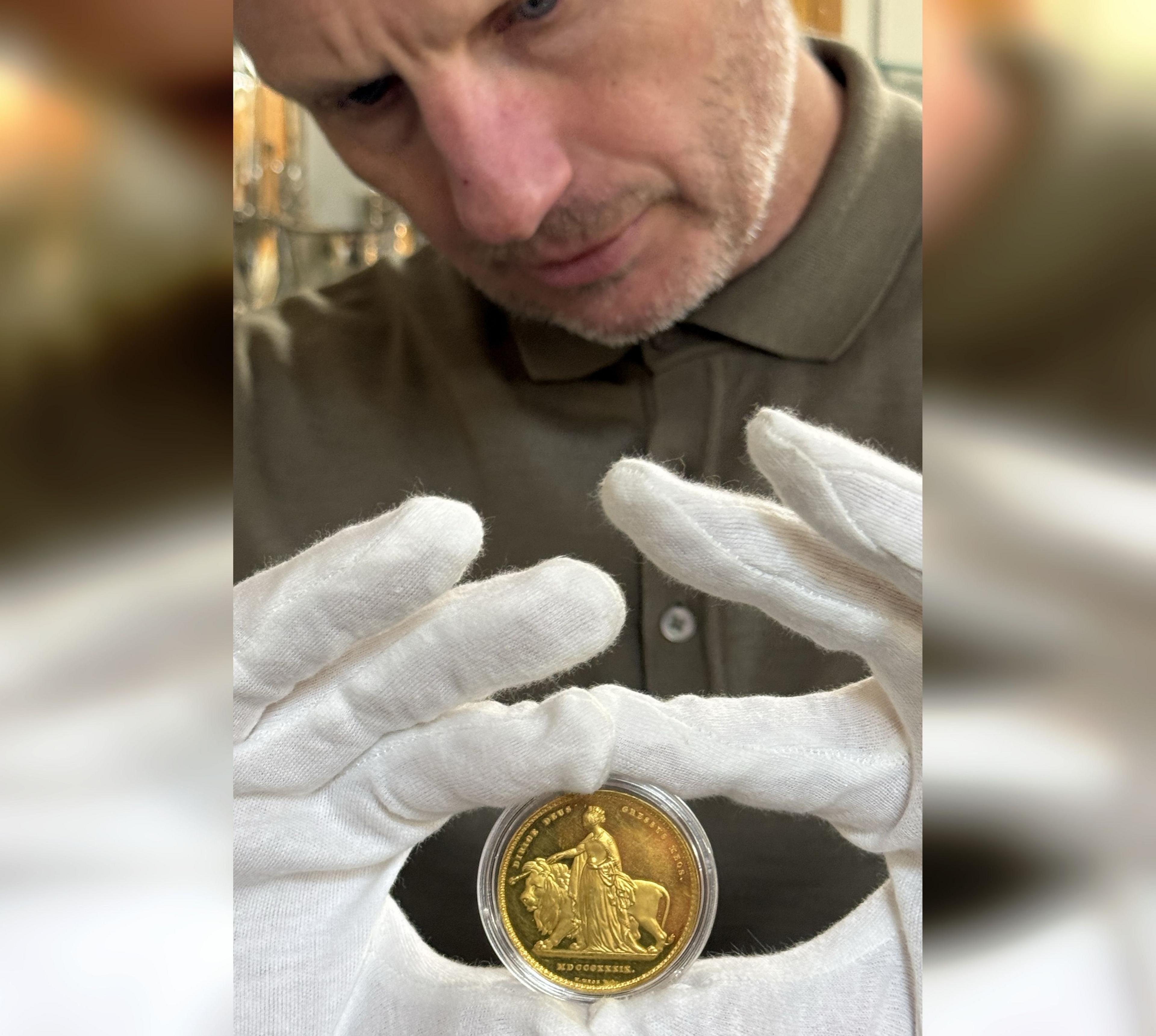 Ben Rogers Jones, gyda'i ddwylo mewn mennyg gwyn, yn dal y darn arian gyda'r dyluniad Una and the Lion tua'r camera