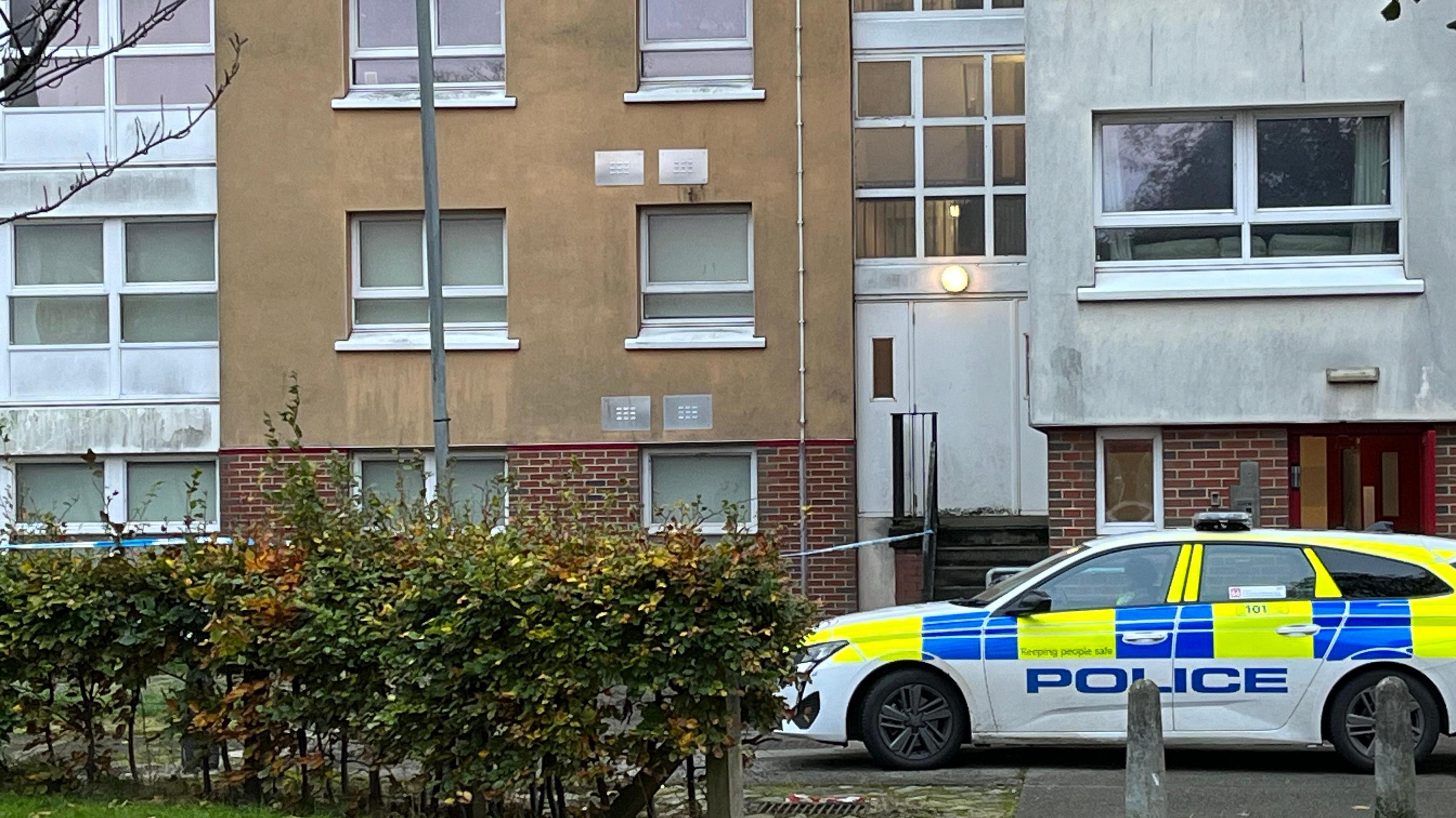 Exterior of a tower block with a police car parked in front and a small area cordoned off by police tape at the front of the building