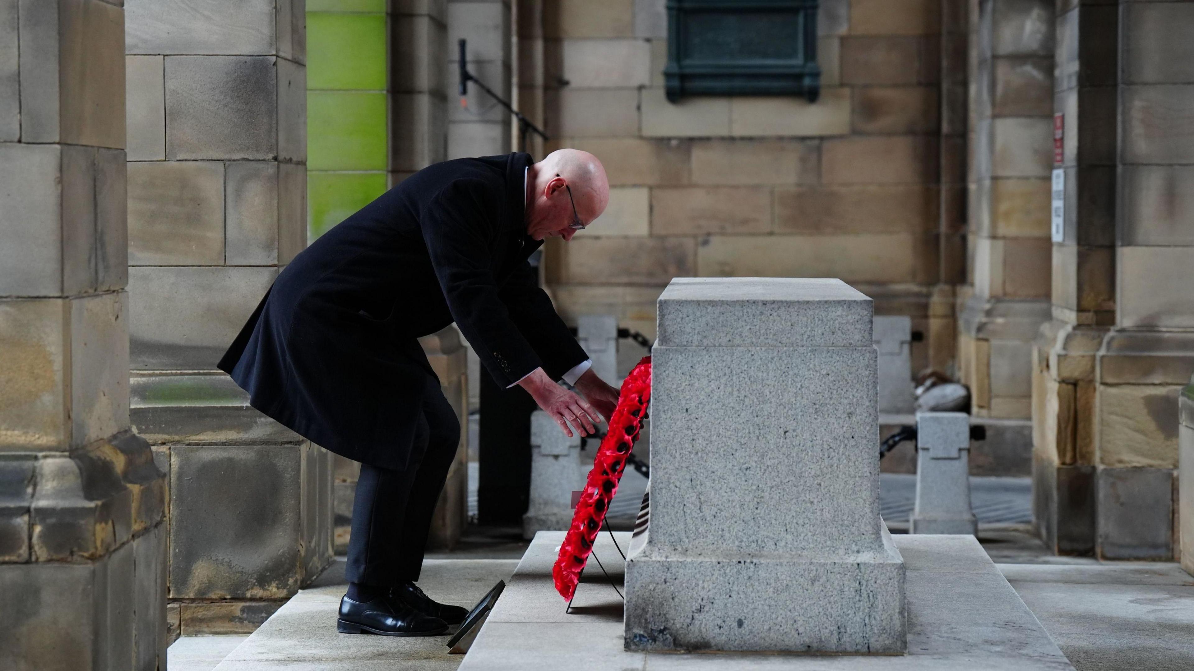 First Minister John Swinney laying a poppy wreath at the Stone of Remembrance in Edinburgh. He is wearing dark clothes, including a long, dark coat.