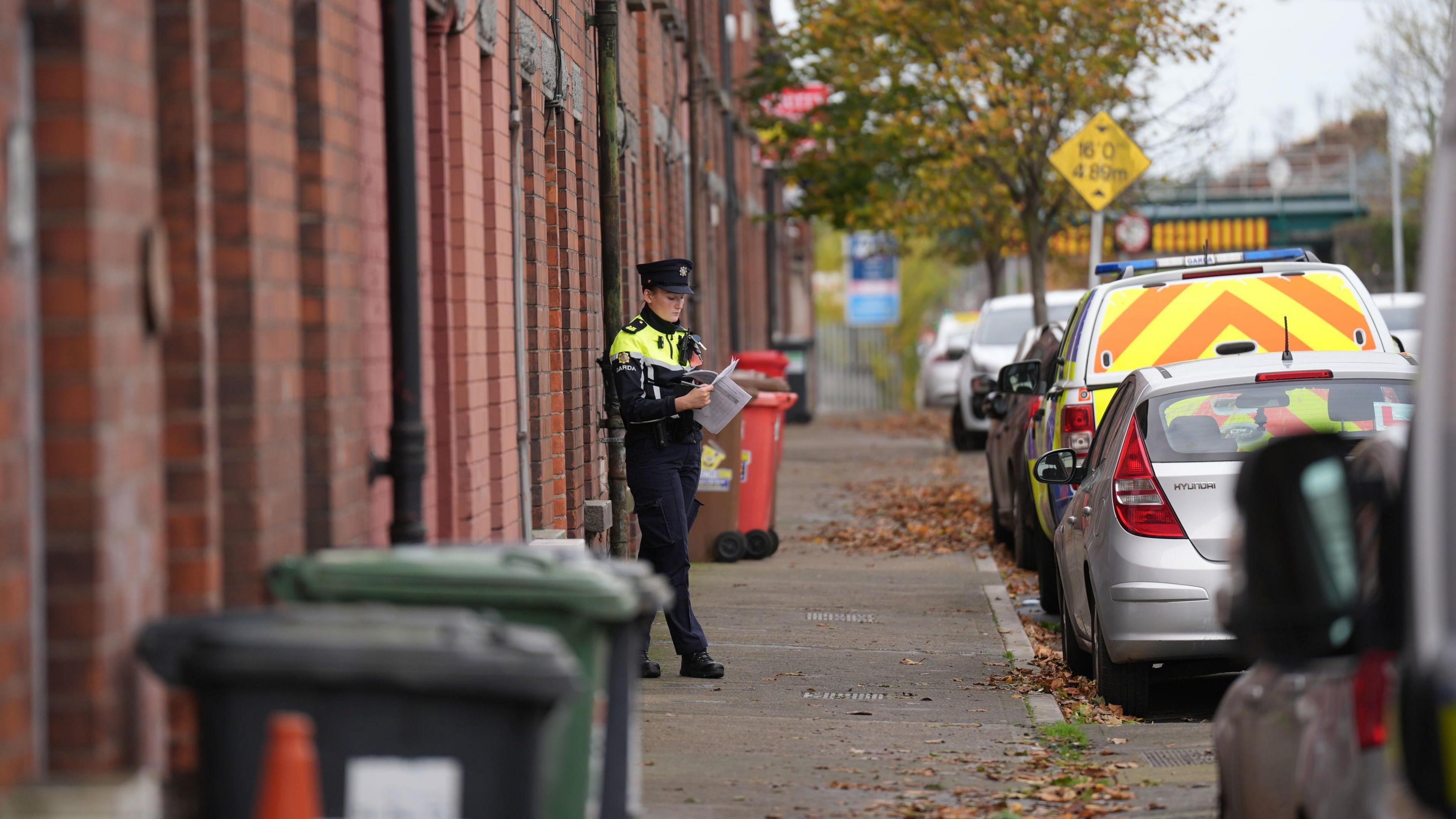 A female garda officer walking out of a house at Emer Terrace, Dundalk. She is wearing full garda uniform and a hat and she is holding documents.