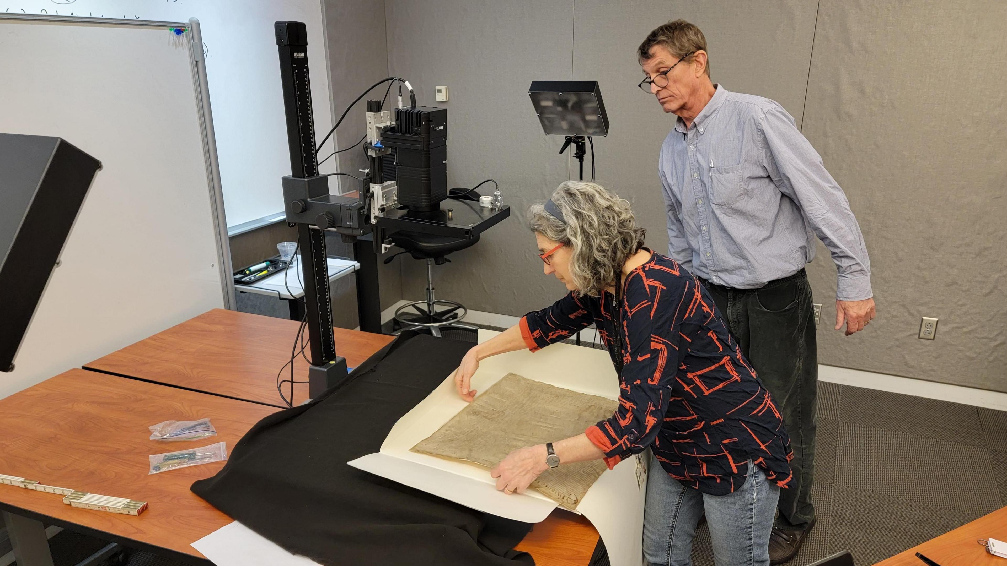 The Harvard Magna Carta placed carefully on a table by a lady wearing glasses. The document is moved under lights in a room where it is tested for its authenticity.