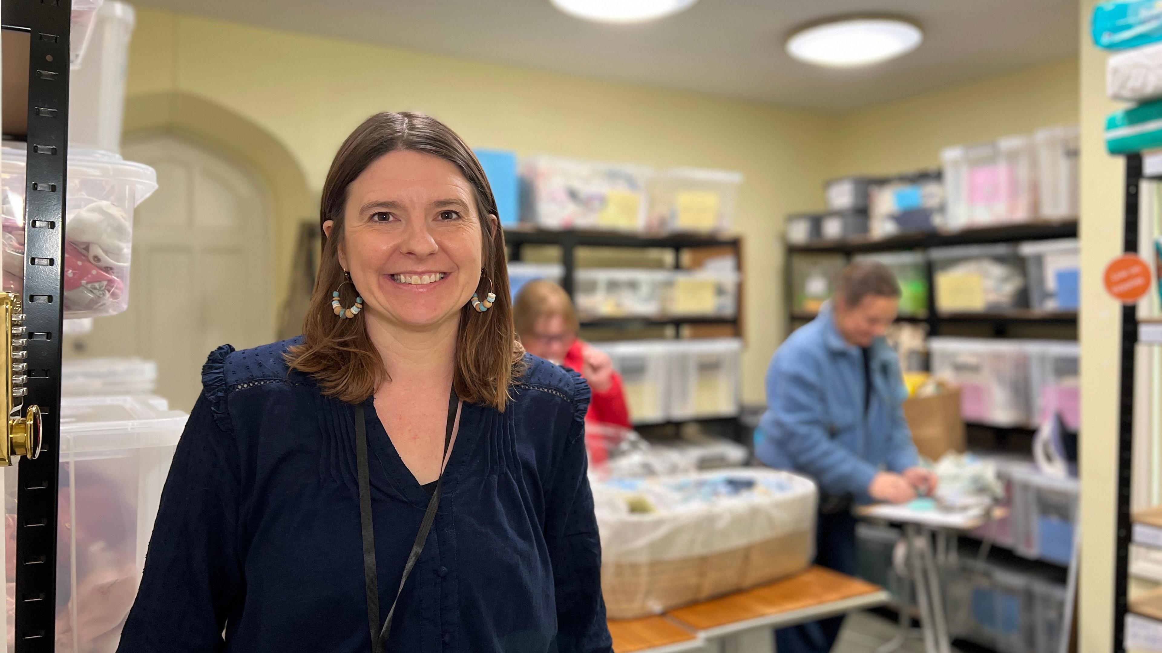 Vicky smiles at the camera as behind her two women are working on packing a Moses basket bed with clothes for a baby. Vicky is wearing a dark blue dress and she has shoulder length brown hair.