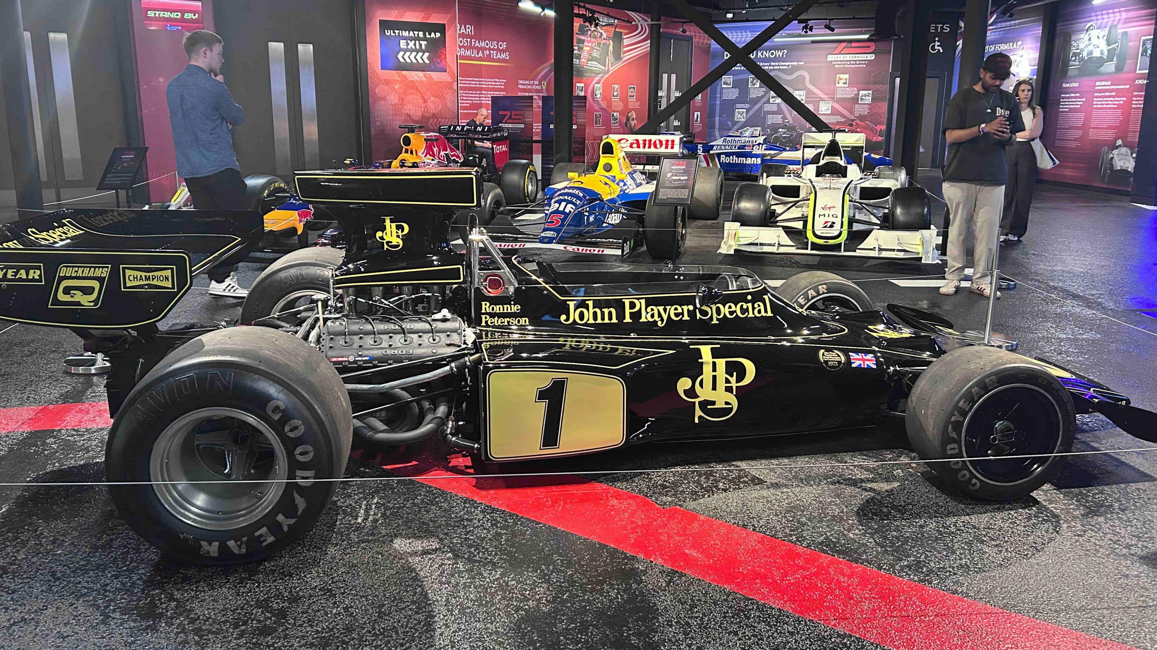 A black racing car with gold writing that says "John Player Special", with a intertwined letter JPS logo and a number one in black with a gold background.
It has large Goodyear tyres. It has Ronnie Peterson's name on the body about where the driver's seat begins. It is parked on a black stage at an exhibition.