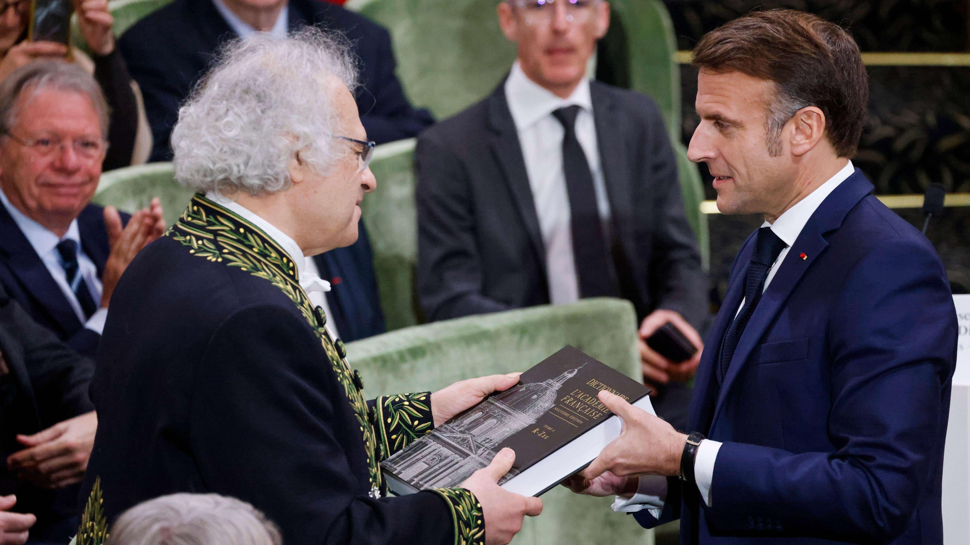 France's President Emmanuel Macron stood on the right receives the 9th edition of the Dictionnaire de l'Academie Francaise from the perpetual secretary of the Academie Francaise, Amin Maalouf, who is stood on the left in a black jack with yellow and green leave-decorated trimming. Macron is wearing a navy blazer and tie.