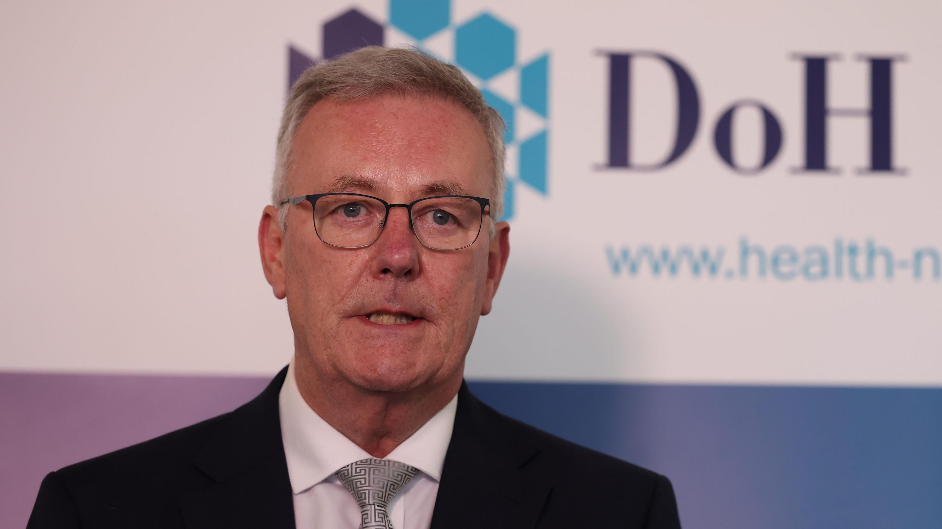 Mike Nesbitt has short grey hair and black framed glasses and is wearing a dark suit jacket, white shirt and silver tie