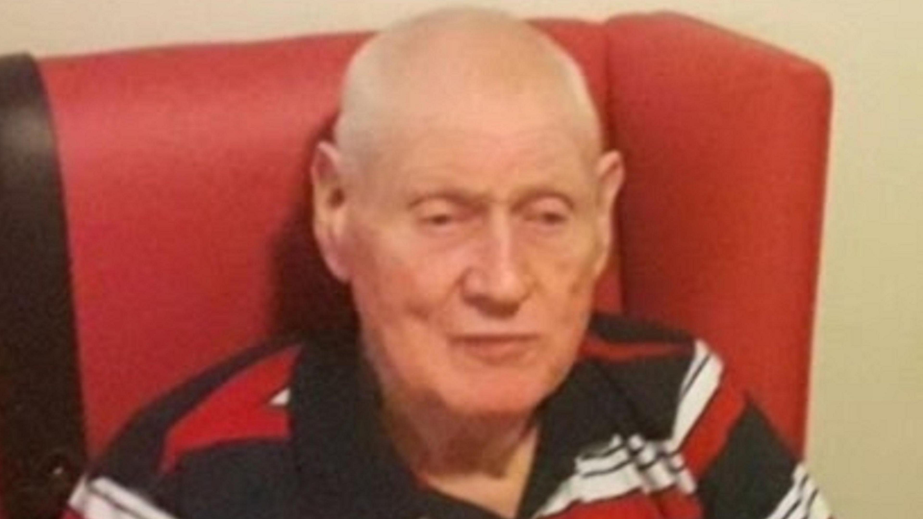 Ivor McQuitty is bald on top with grey hair to the sides. He is wearing a red, black and white polo shirt and sitting in a red chair.