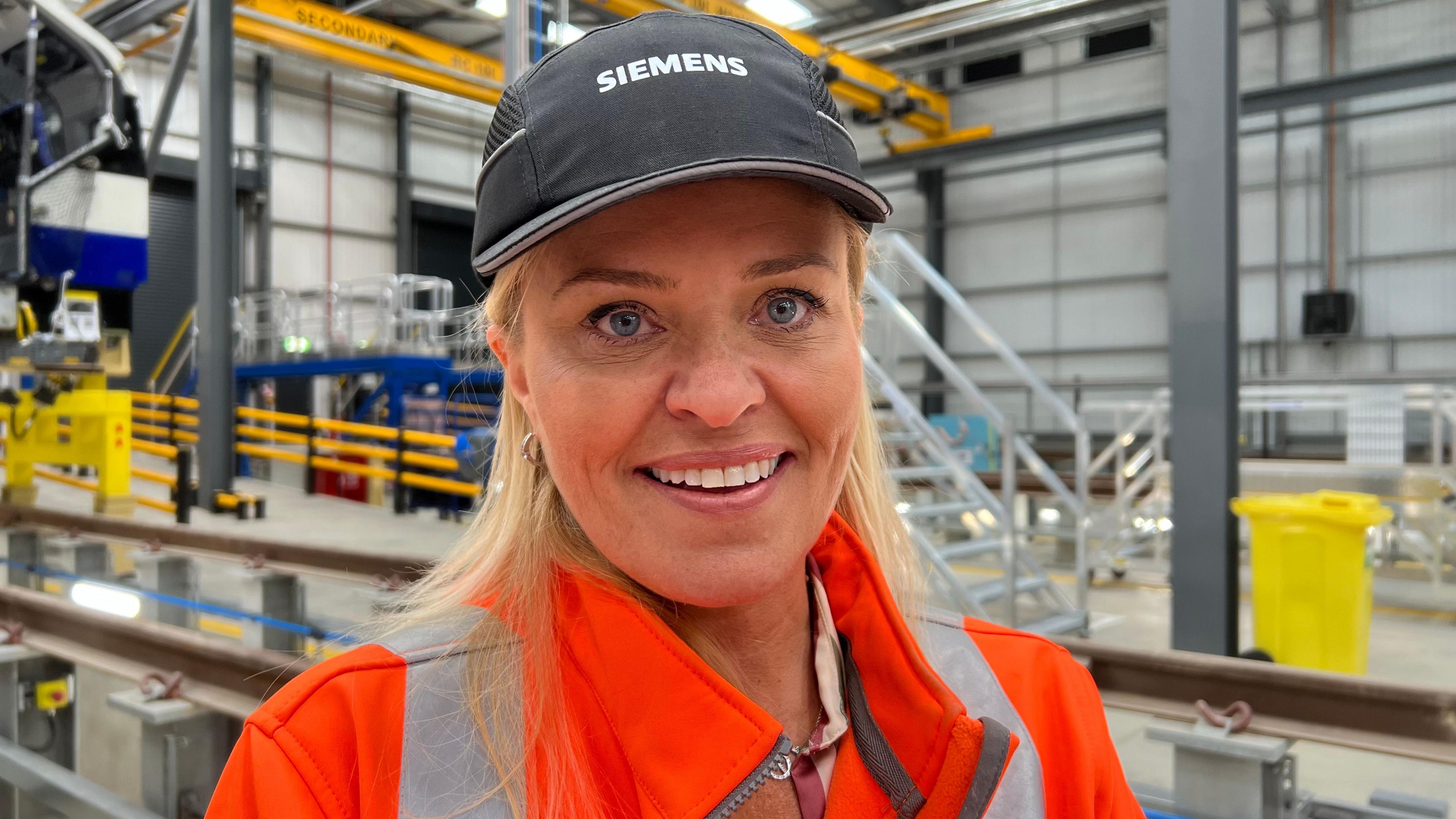 Natalie is wearing an orange hi-vis vest and a black Siemens' cap over her long blonde hair. She is smiling at the camera on the factory floor