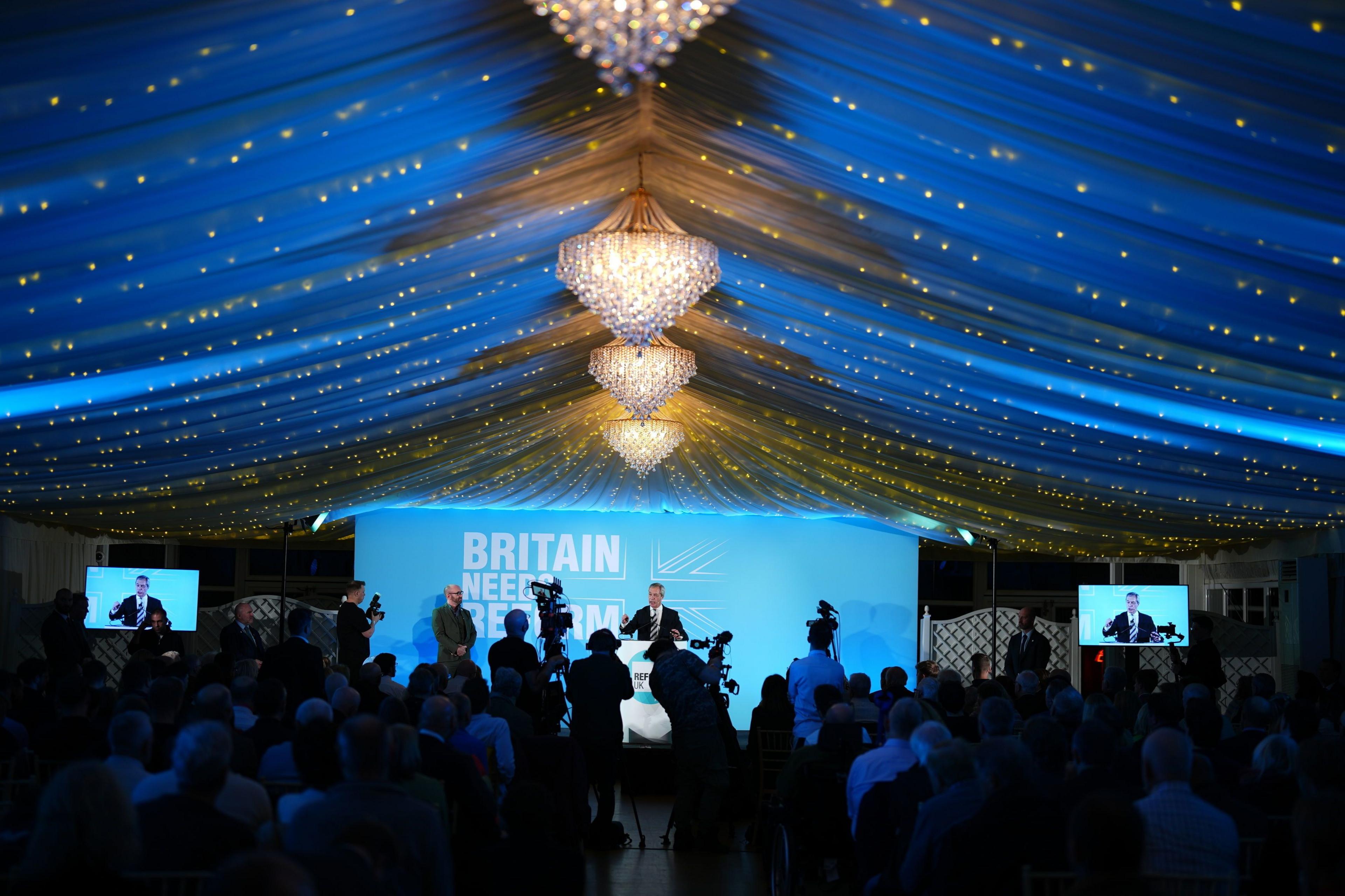 Nigel Farage on stage speaking under a chandelier during a Reform UK conference. The wide angle shot shows a line of photographers next to the stage with rows of seated audicnce members behind